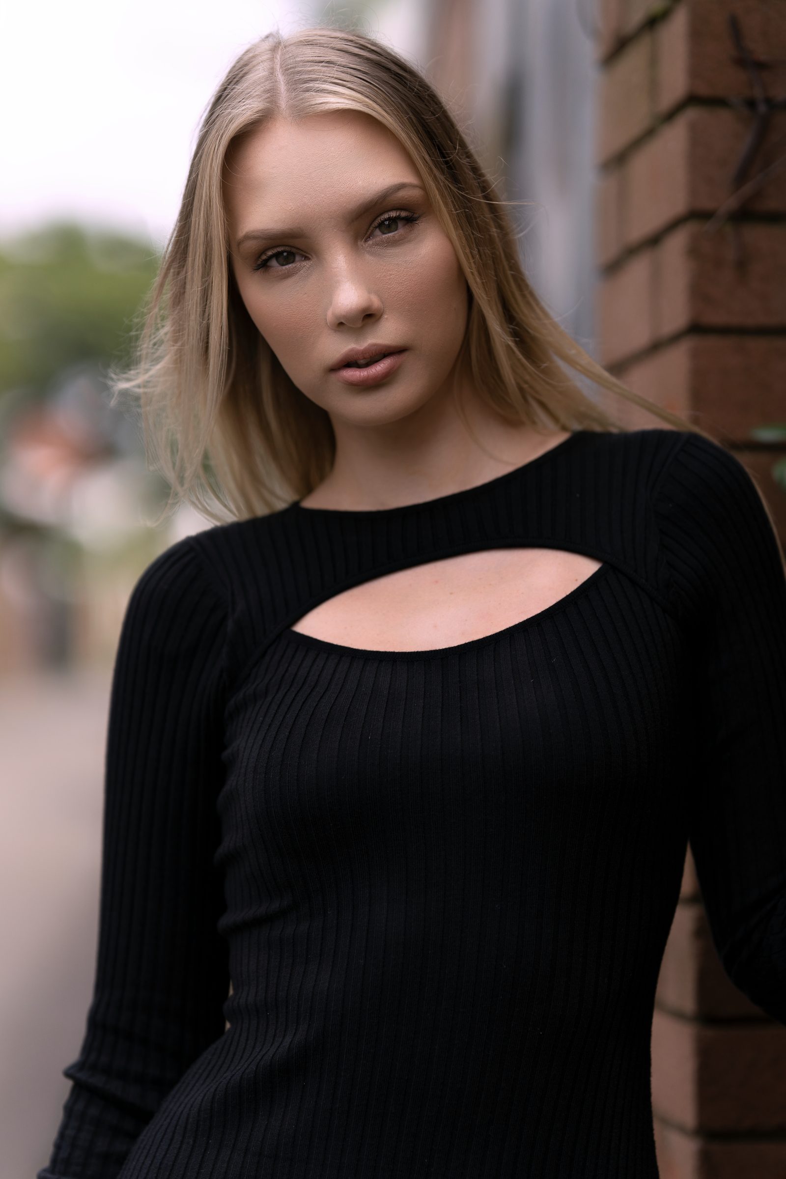 A close portrait of a woman in a black ribbed top with a keyhole cut-out, leaning against a brick wall