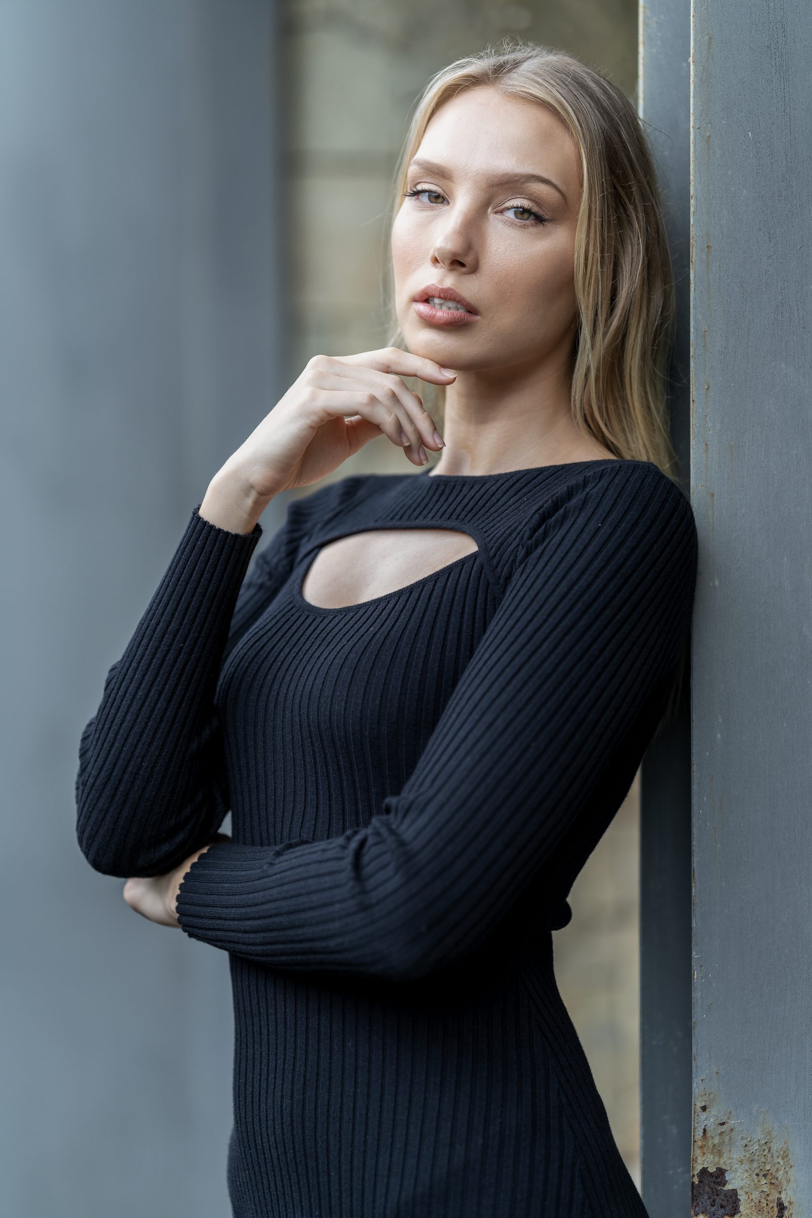 A woman in a black ribbed cut-out dress rests a hand near her chin, leaning against a grey steel column