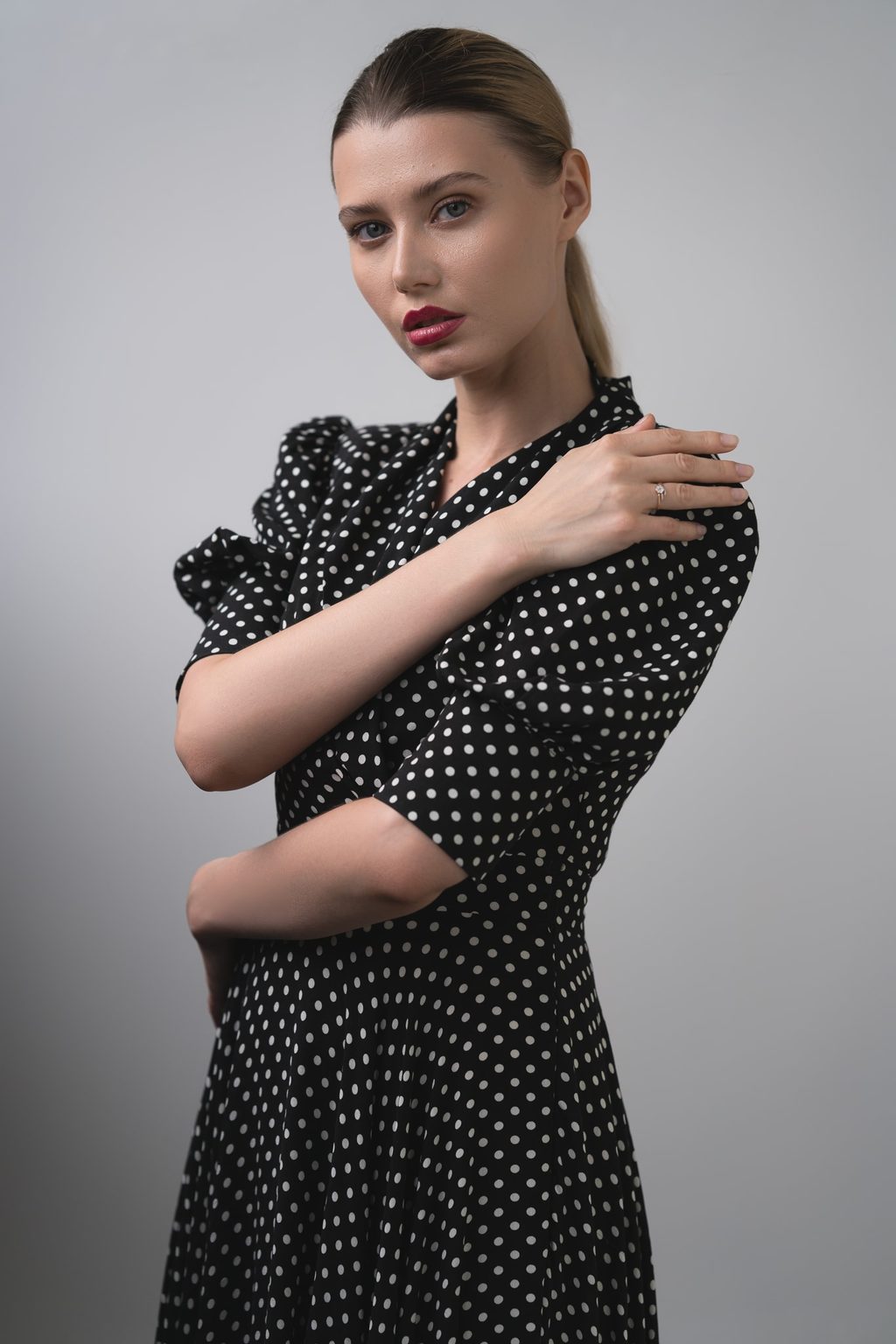 A woman in a black polka dot dress with puffed sleeves rests one hand on her shoulder, posing against a plain grey backdrop