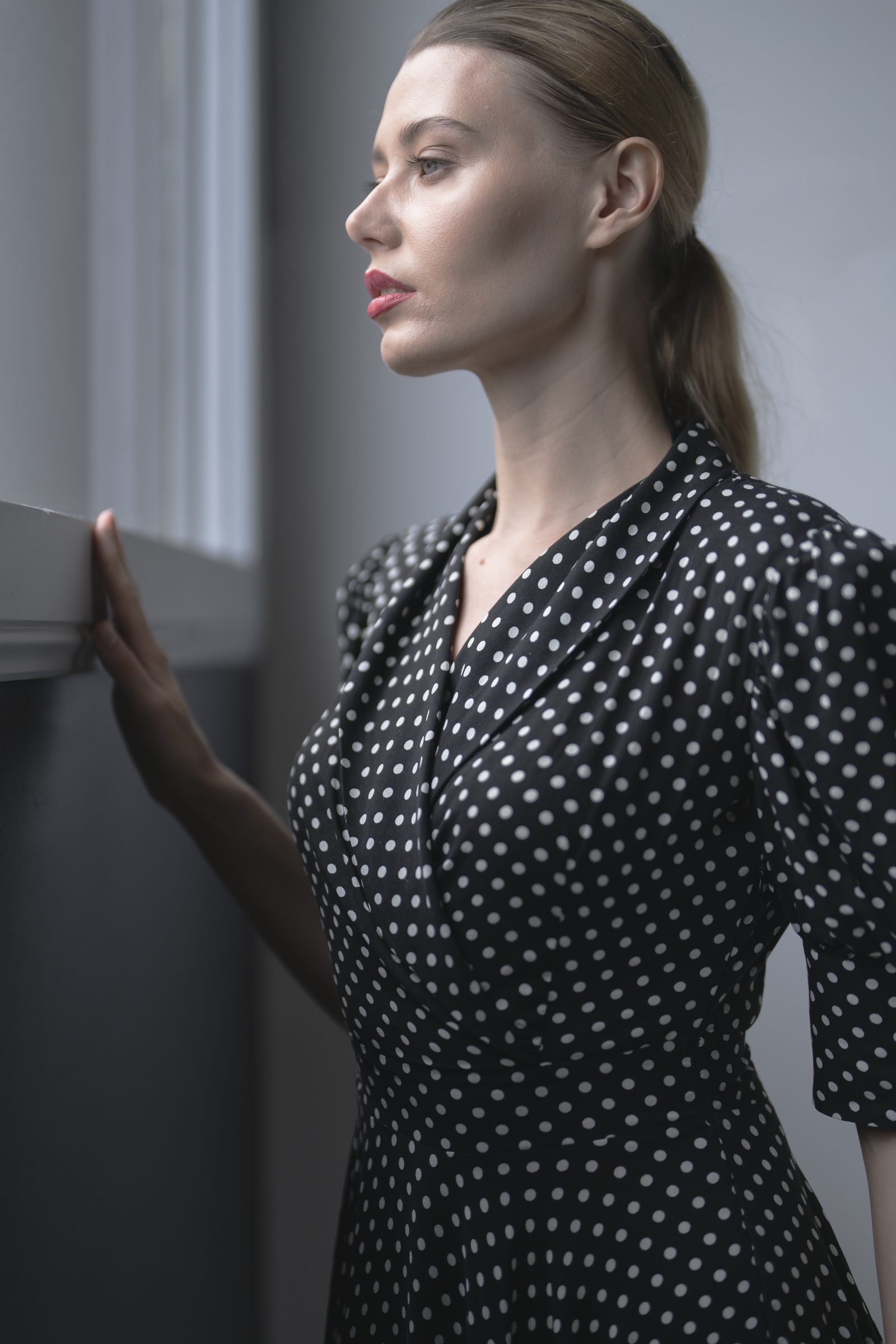 A woman in a black and white polka dot dress stands in profile at a window with one hand on the sill