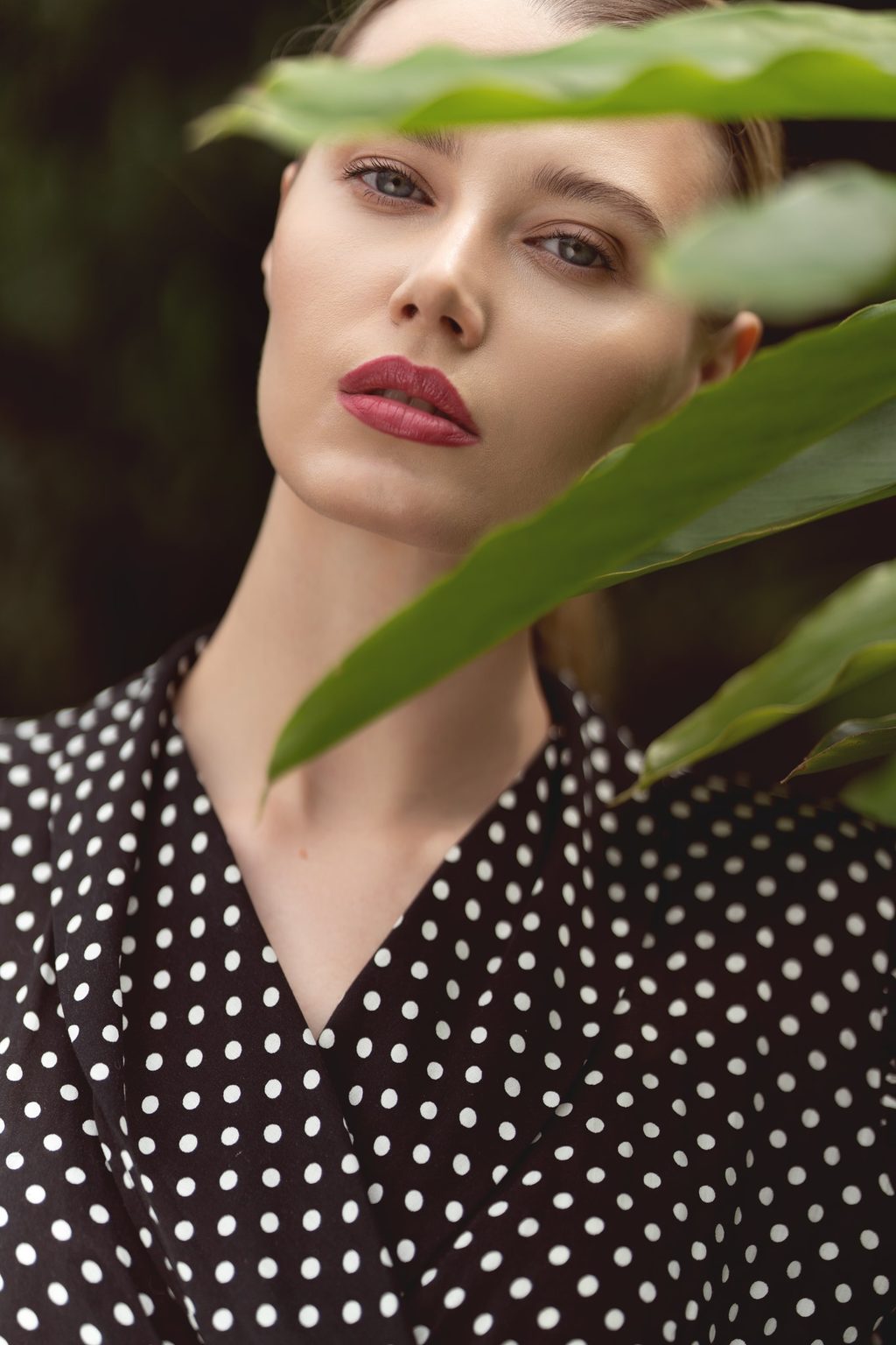A woman in a black and white polka dot top peers past a large green leaf, her lips in deep red against a dark background