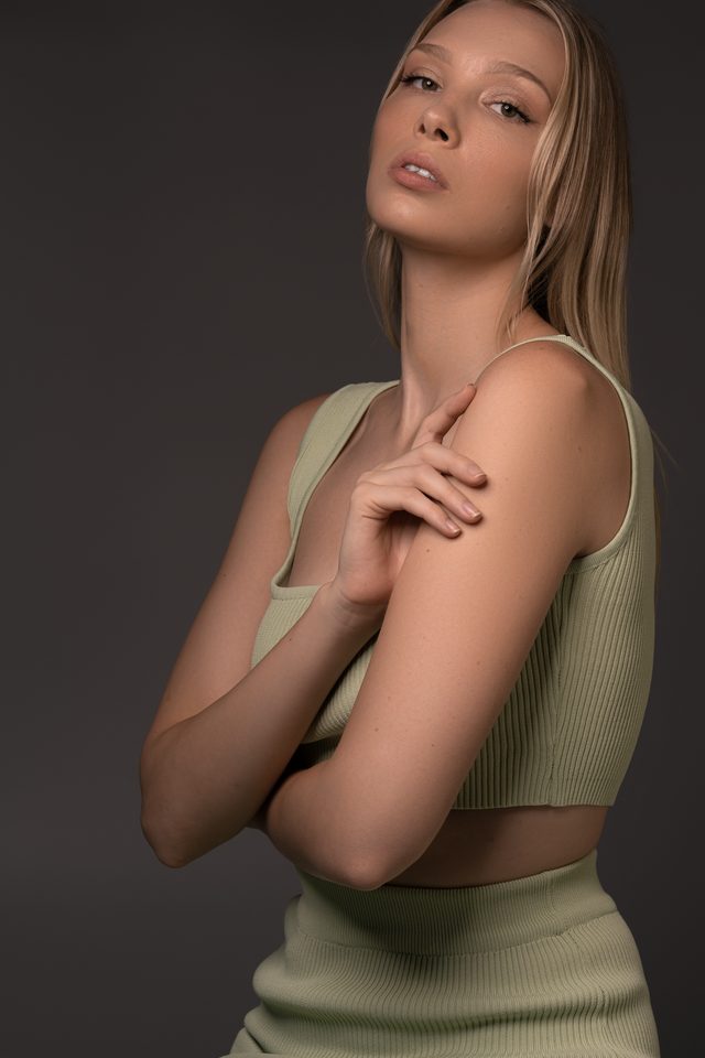 A woman in a sage green ribbed crop top and skirt turns to camera with one hand resting on her upper arm