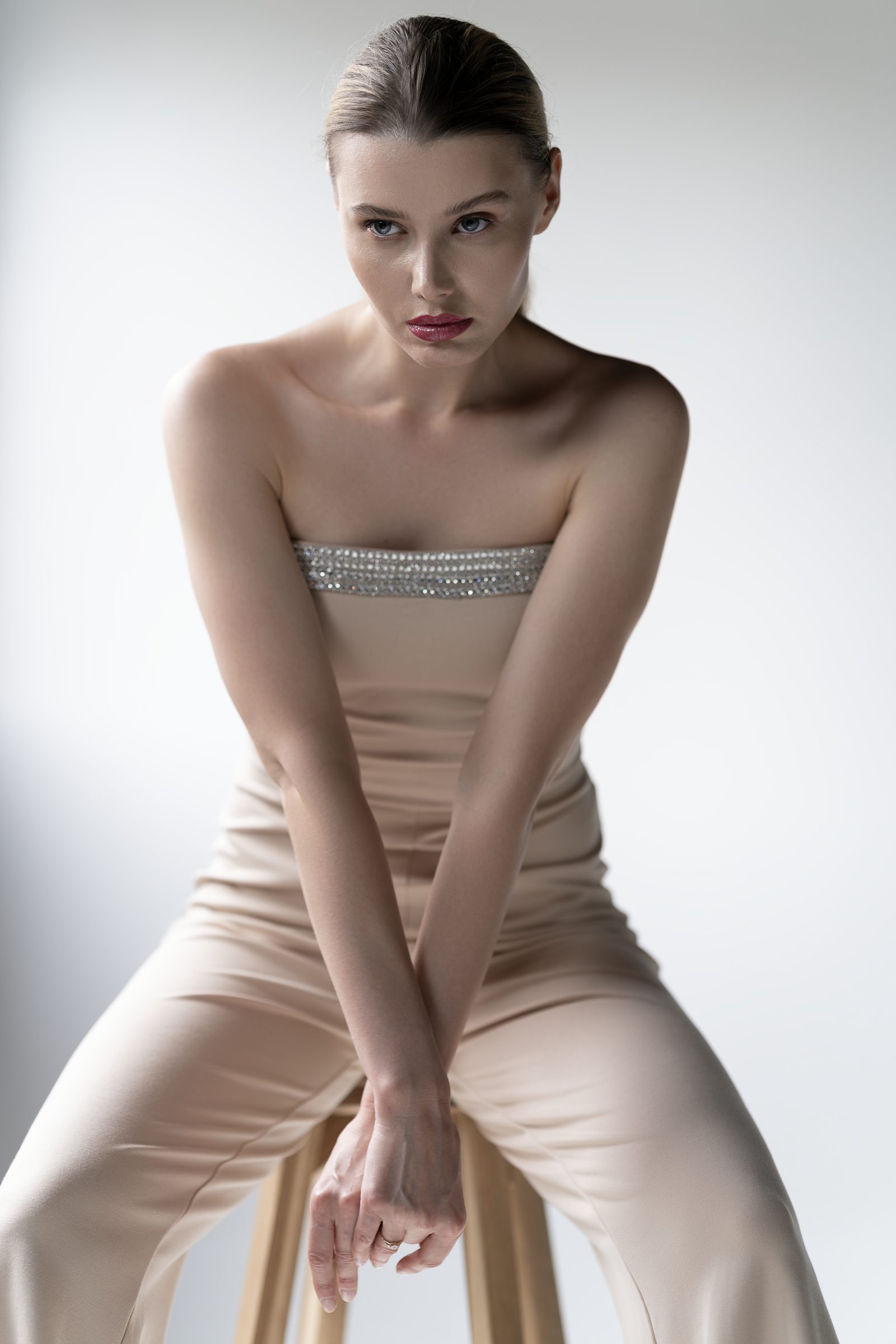 A woman in a strapless champagne-coloured jumpsuit sits with arms crossed on her knees, against a bright white studio background
