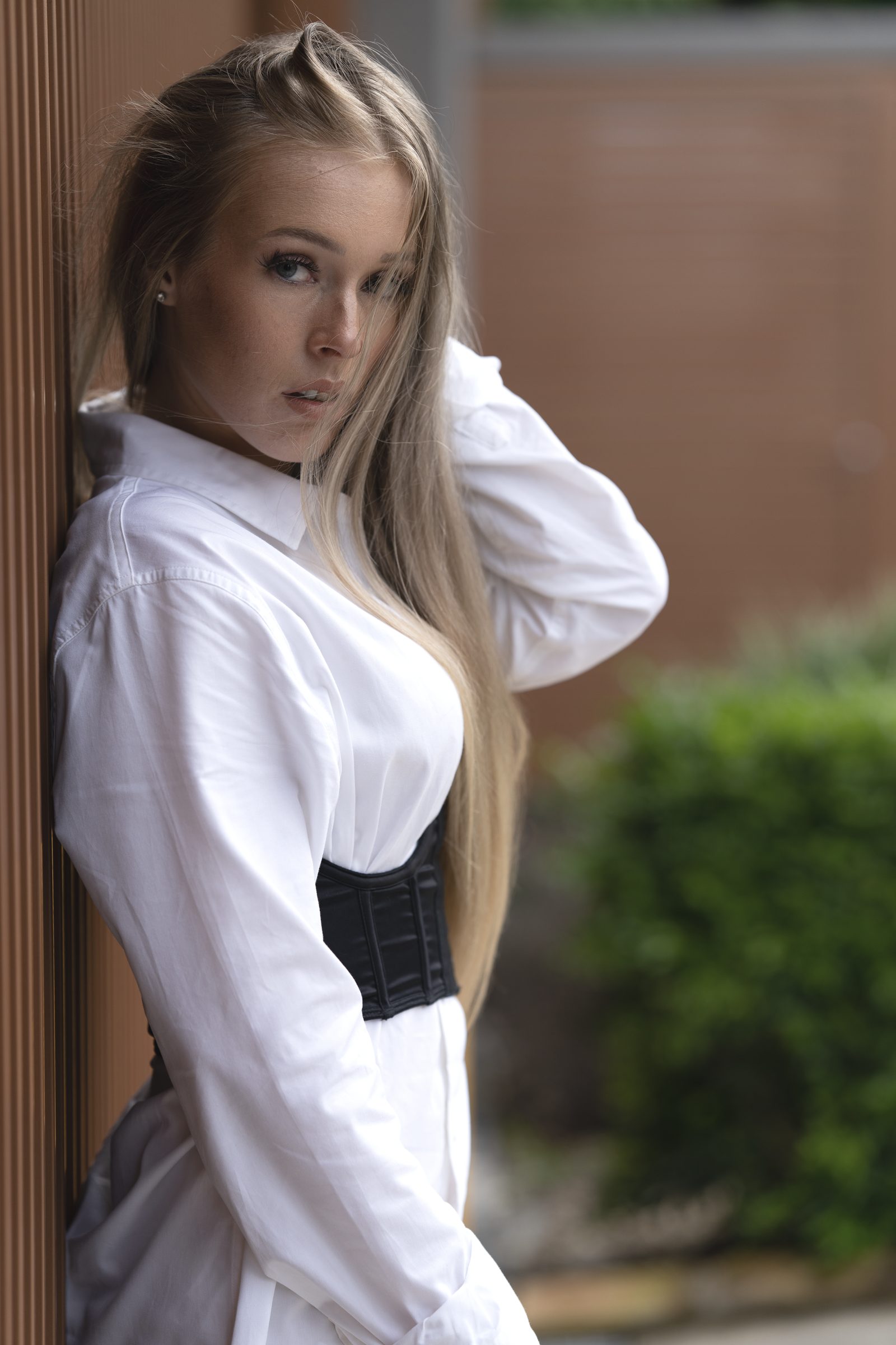 A woman in an oversized white shirt cinched with a black corset belt leans against a timber post, one hand behind her head