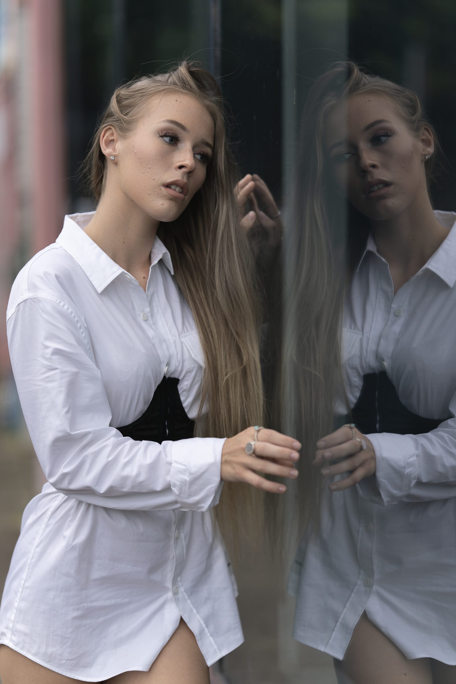 A woman in a white shirt dress stands against a glass shopfront, her reflection doubling her alongside