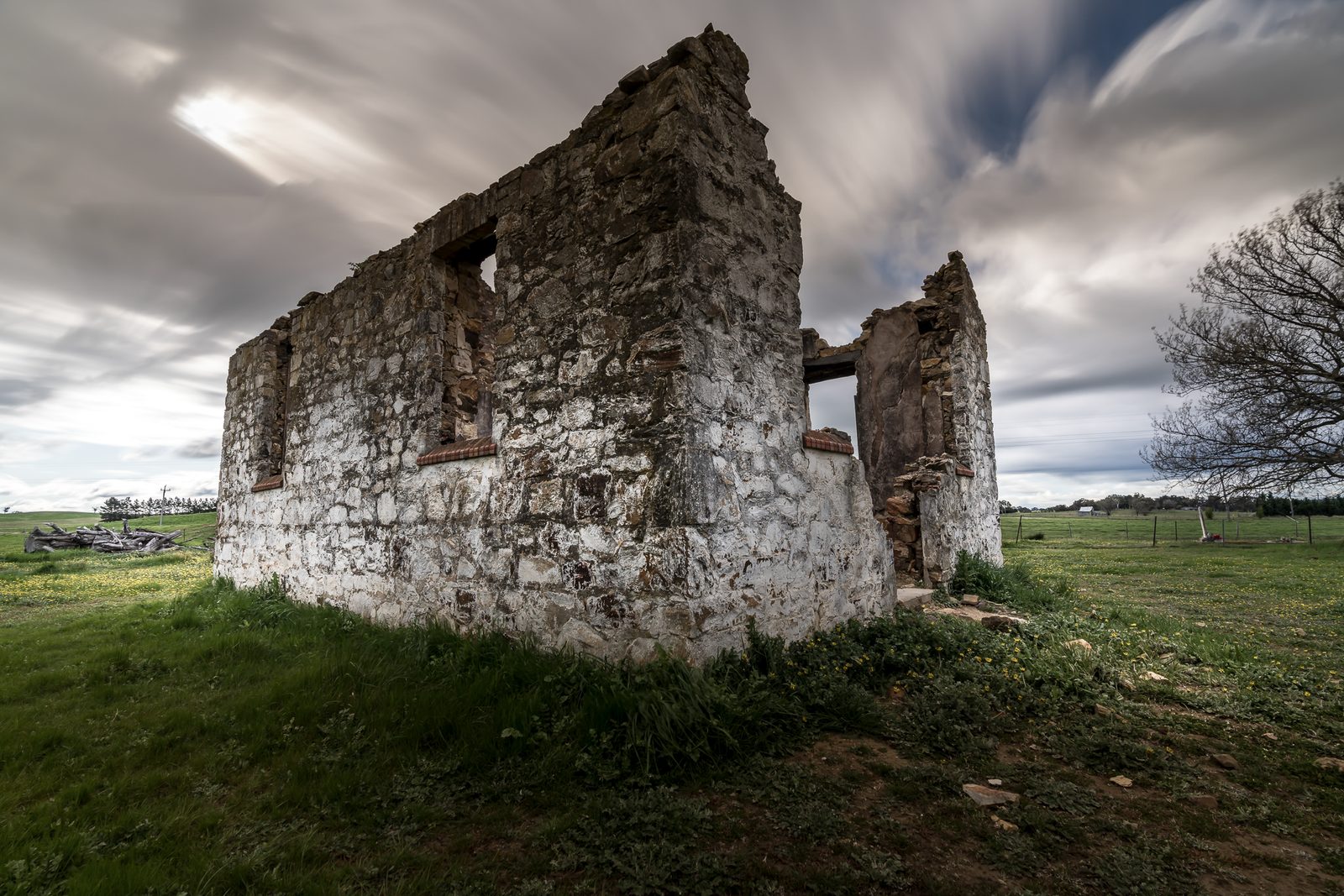A roofless stone farmhouse stands in a green paddock, its whitewashed walls open to fast-moving cloud above.