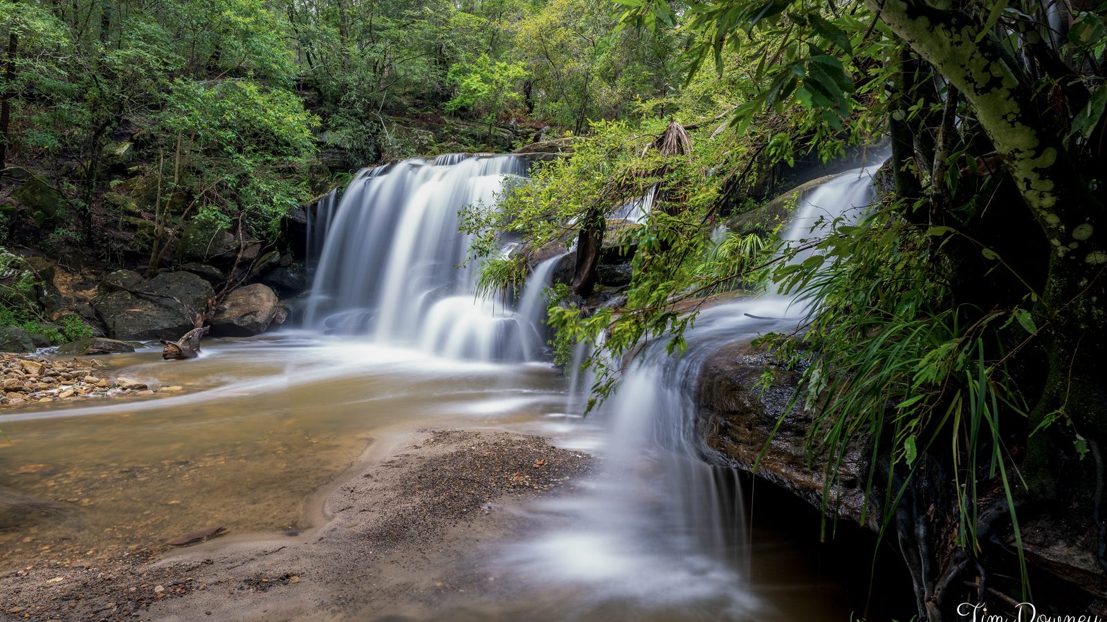 A waterfall slides in silk sheets over sandstone ledges into a shallow pool, ferns and green scrub crowding the banks.