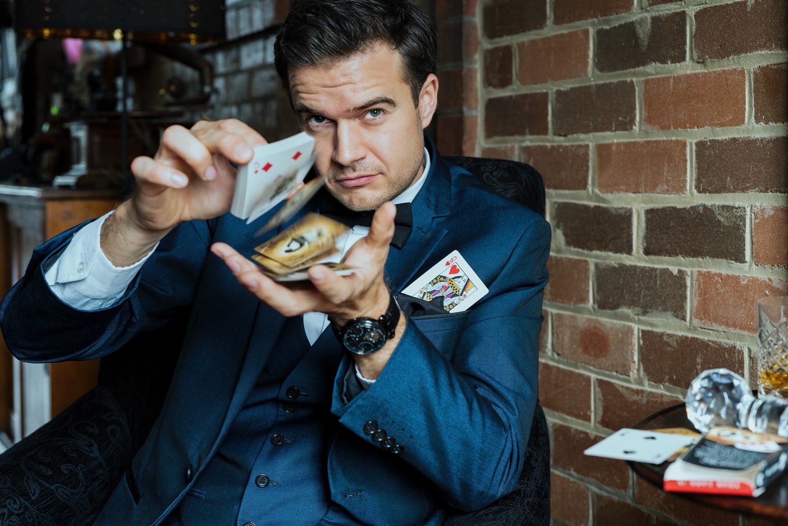 A man in a navy three-piece suit and bow tie fans a deck of playing cards, a queen of hearts tucked in his breast pocket