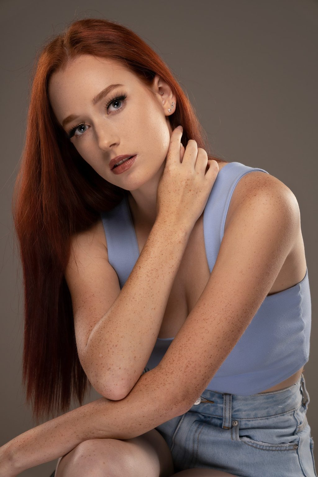 A freckled woman with long red hair rests her hand near her neck, wearing a pale blue top against a grey studio backdrop