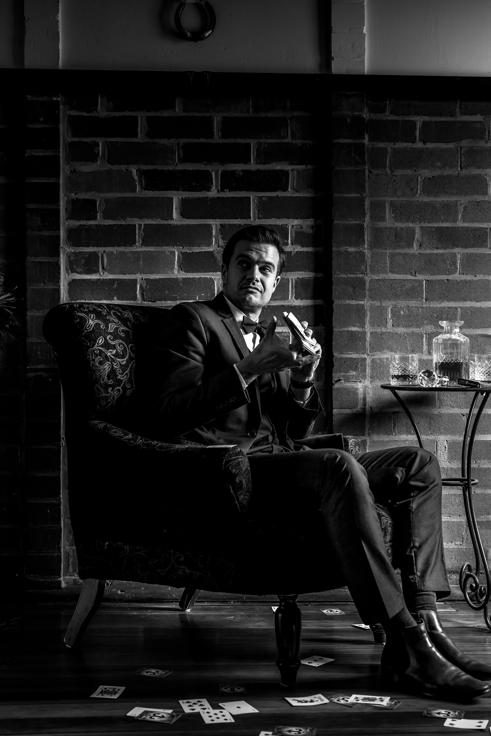 In black and white, a man in a three-piece suit and bow tie sits in a wing chair holding playing cards, more cards scattered across the floor