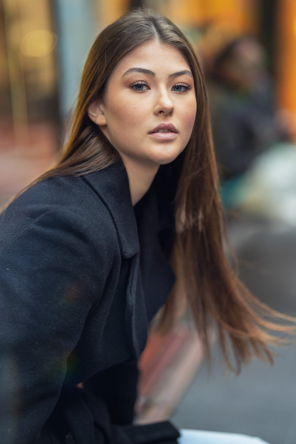 A woman with long straight hair looks over her shoulder in a black coat on a blurred city street with reflections
