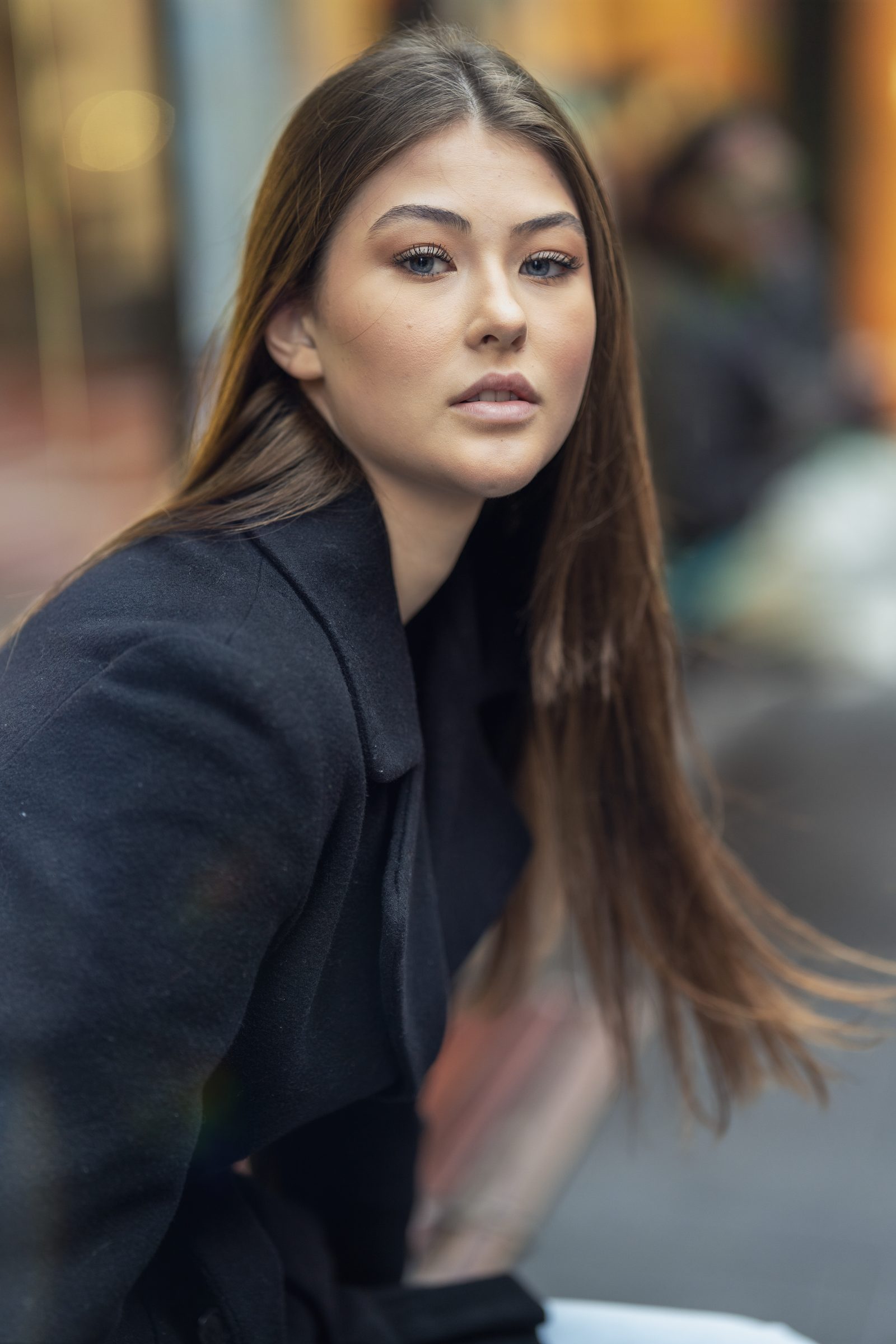 A woman with long straight hair looks over her shoulder in a black coat on a blurred city street with reflections
