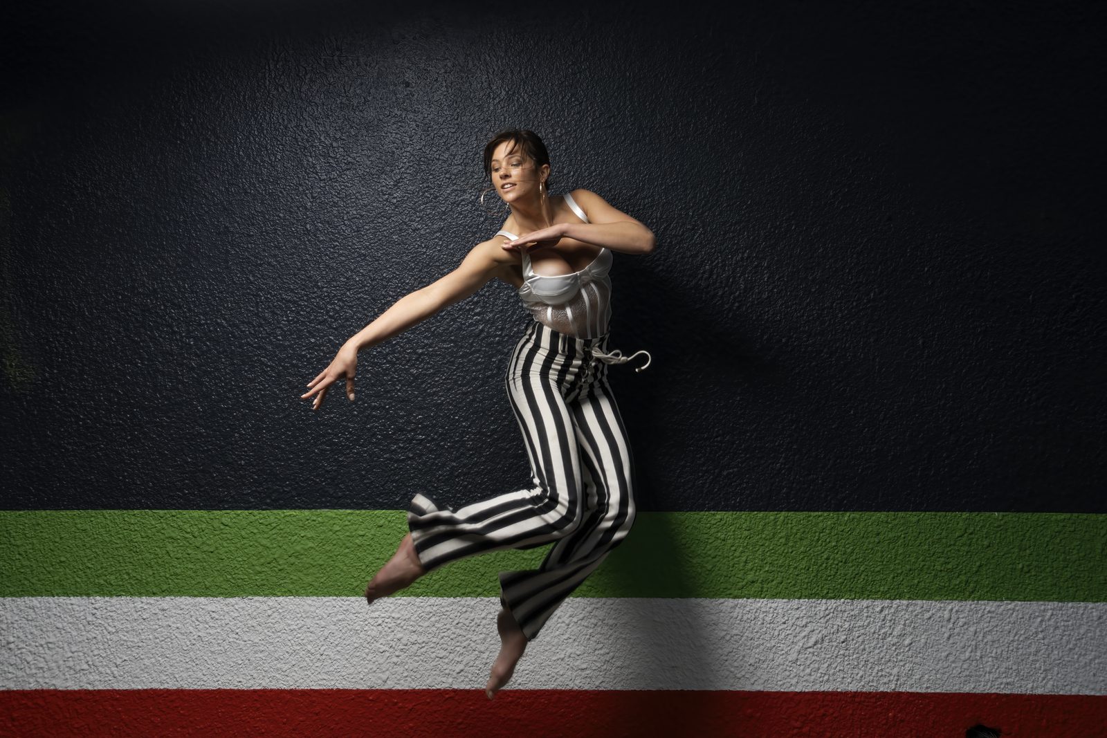 A woman in a white bustier and black and white striped trousers hangs mid-air against a wall banded in green, white and red