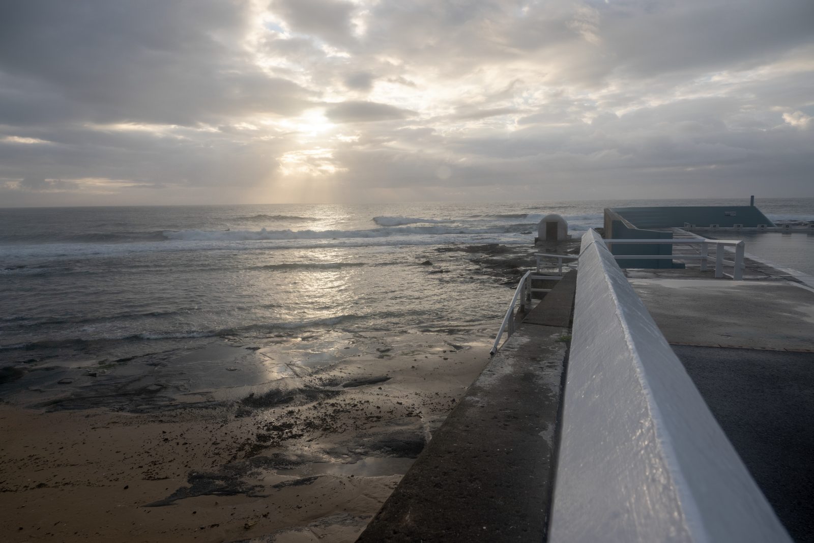 The concrete wall of an ocean pool runs out toward a rock shelf at dawn, sun breaking through cloud over the surf.