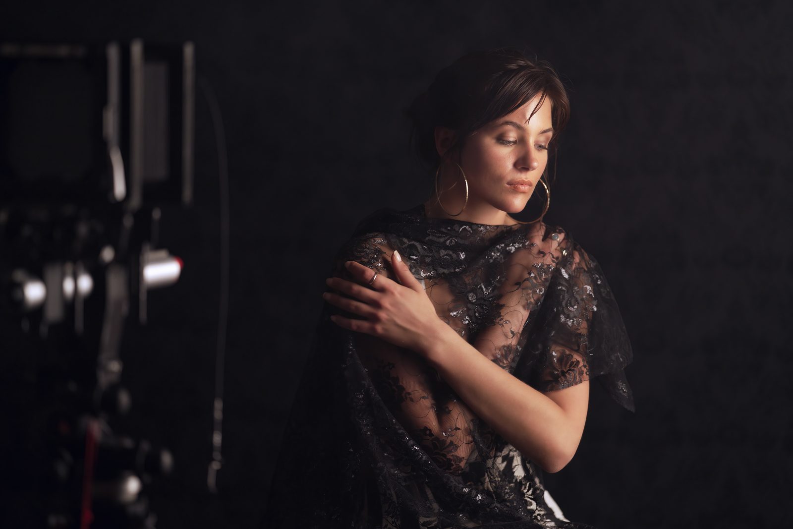 A woman in black sequinned lace over a white top holds it closed at her shoulder, a studio light just in frame