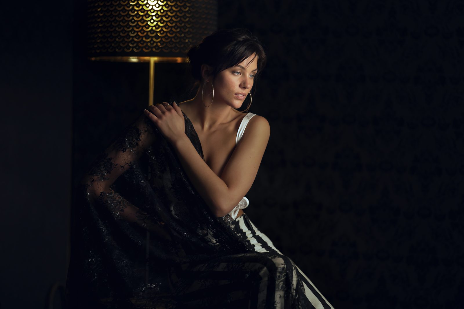 A woman in black lace and a white top sits beside a perforated gold floor lamp, a dark patterned wall behind her