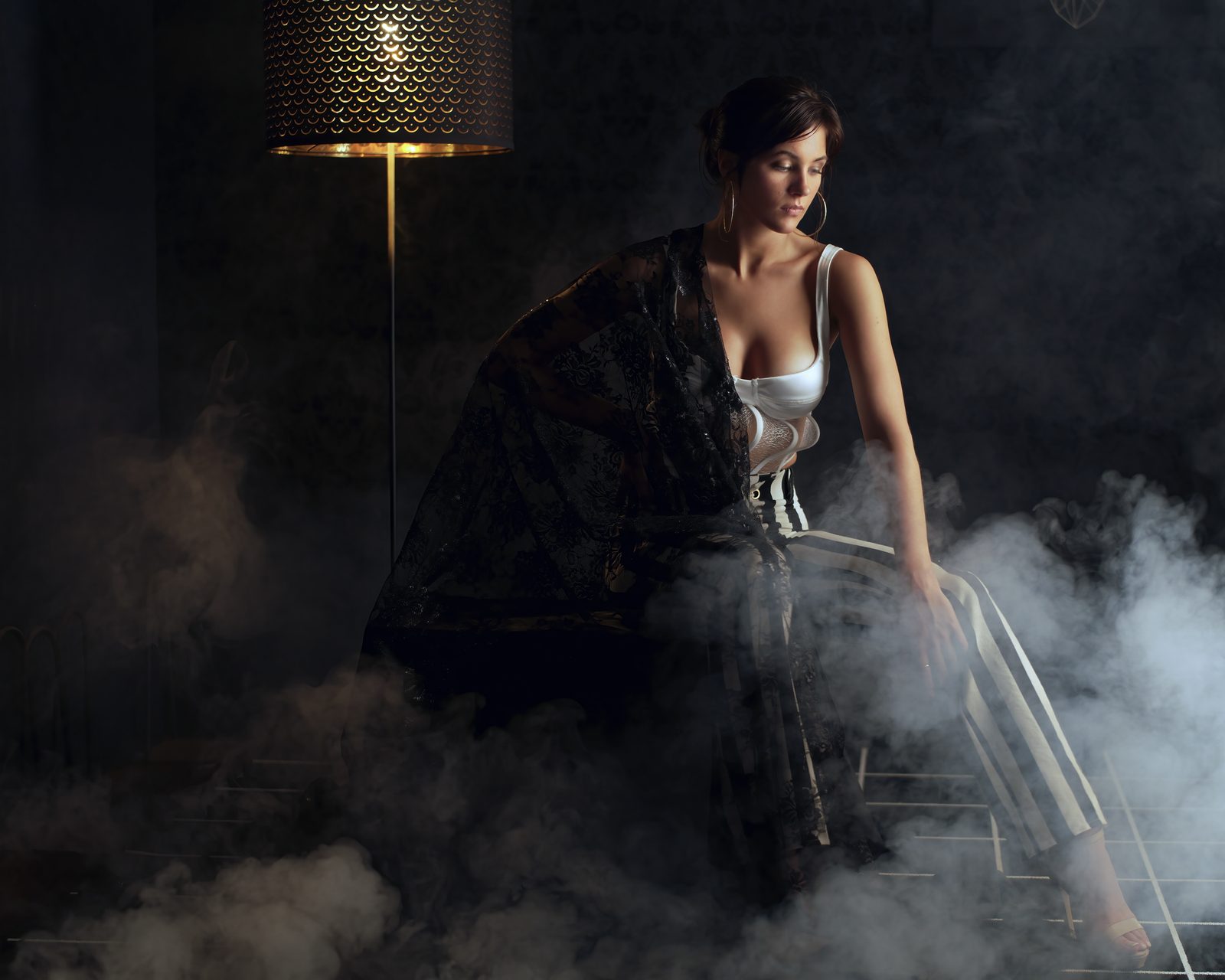 A woman in black lace over white sits in low drifting smoke beside a gold floor lamp, looking down and away