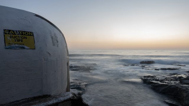 A faded caution sign on the curved white wall of an ocean pool, long-exposure surf washing the rock shelf beyond at dusk.