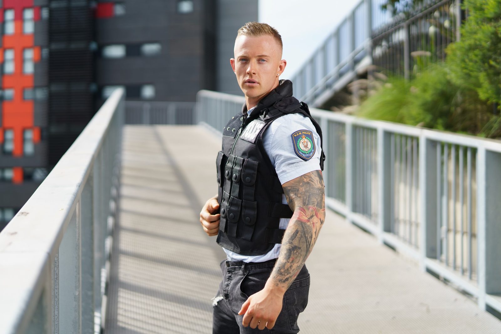 A man in a police-style tactical vest and ripped jeans stands on an elevated walkway, a large tattoo visible on his forearm
