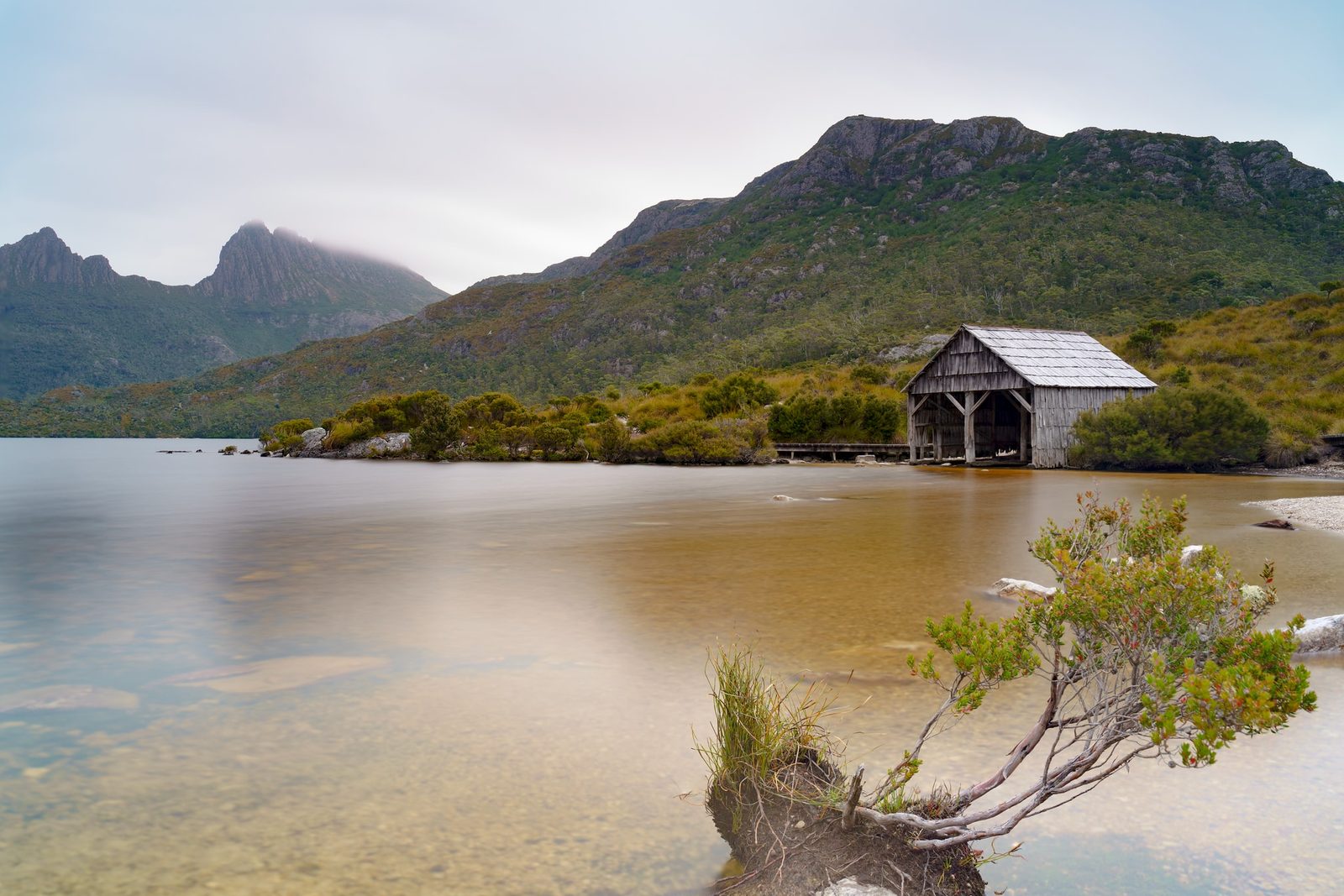 A weathered timber boat shed sits on the shore of a still lake, with a jagged mountain range rising behind.