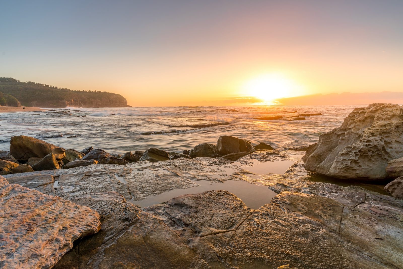 Sunset breaks over a headland as waves wash across a flat sandstone shelf pooled with water in the foreground.