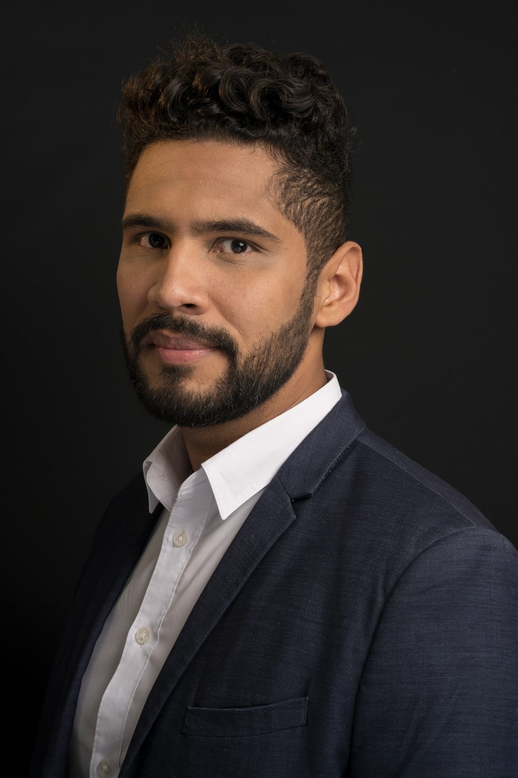 A bearded man in a navy suit and white shirt poses for a headshot, turned slightly to camera against a plain black background