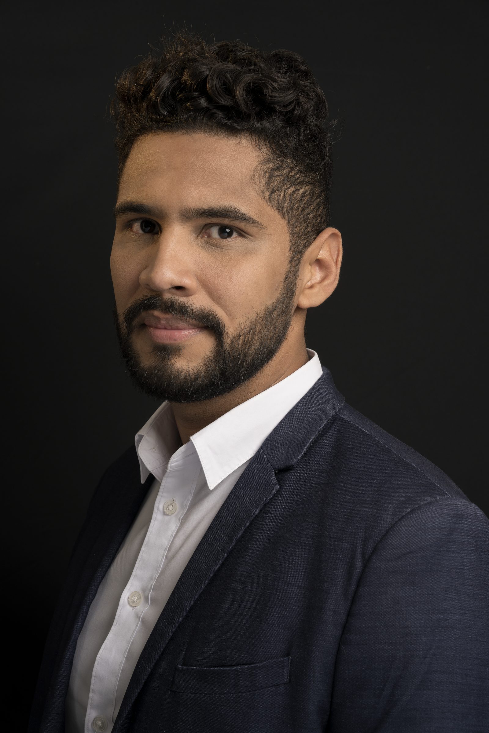 A bearded man in a navy suit and white shirt poses for a headshot, turned slightly to camera against a plain black background
