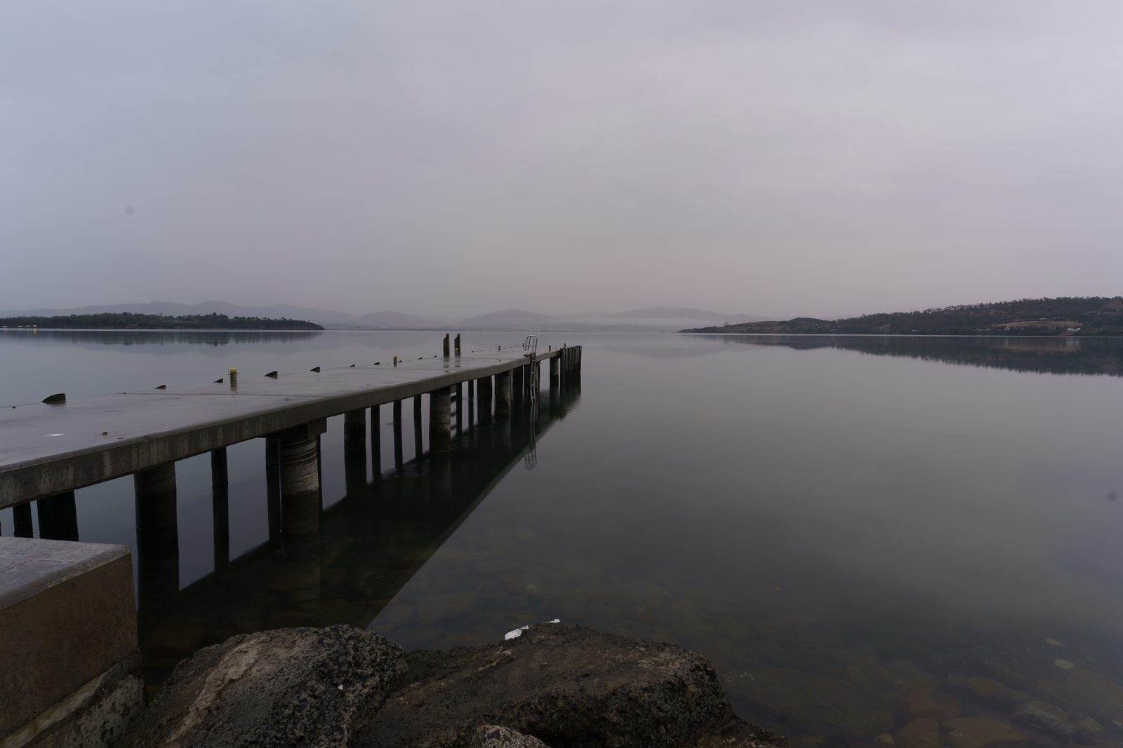 A concrete jetty runs out into glassy grey water toward two distant figures, low hills fading in the haze.