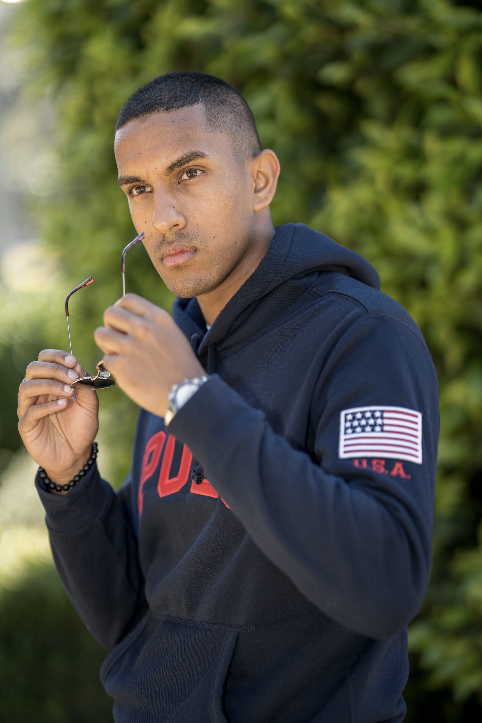 A man in a navy hoodie holds a pair of sunglasses up near his mouth, a dark green hedge behind him