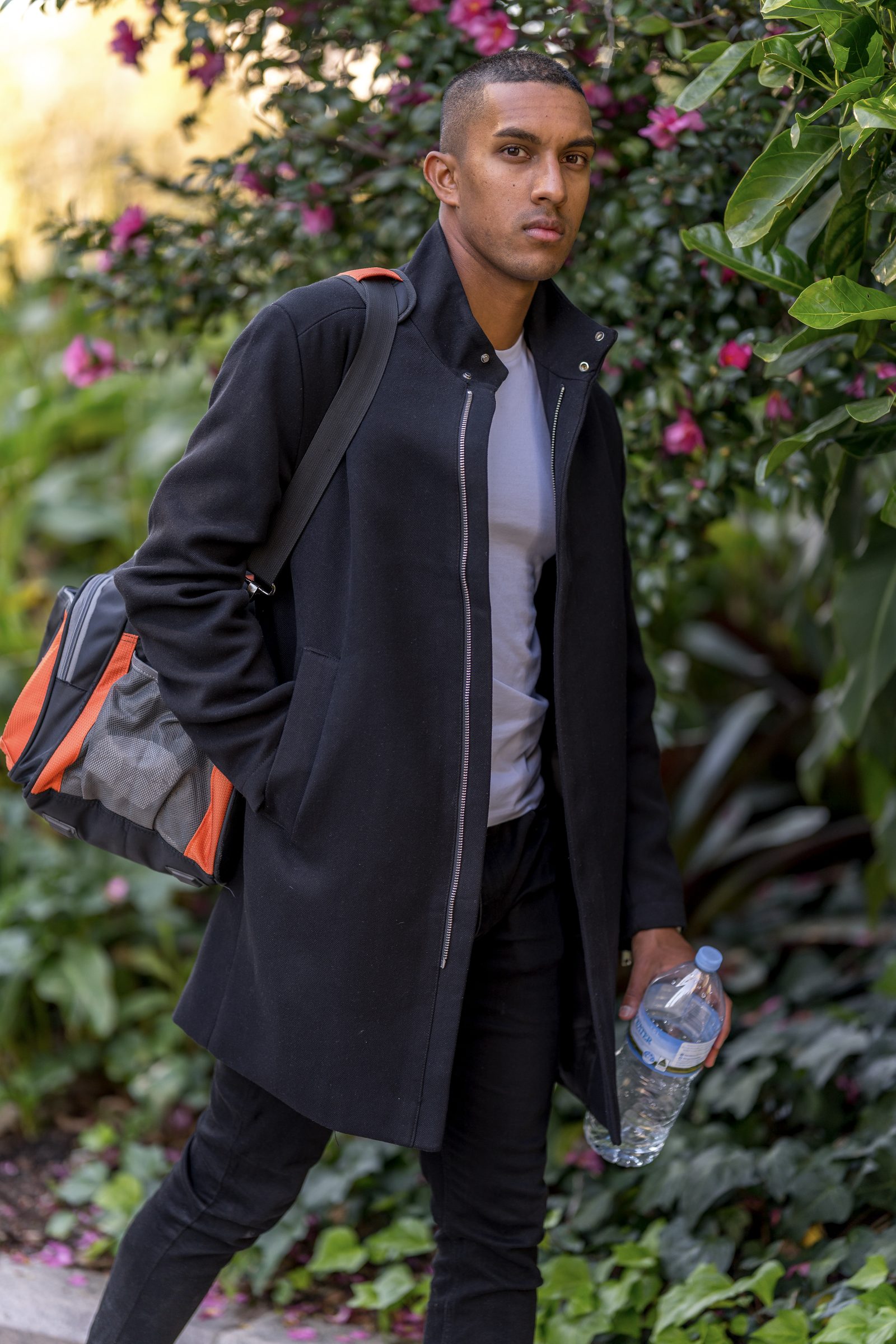 A man in a long black coat carries a duffel bag over one shoulder and a water bottle in hand, standing before flowering shrubs