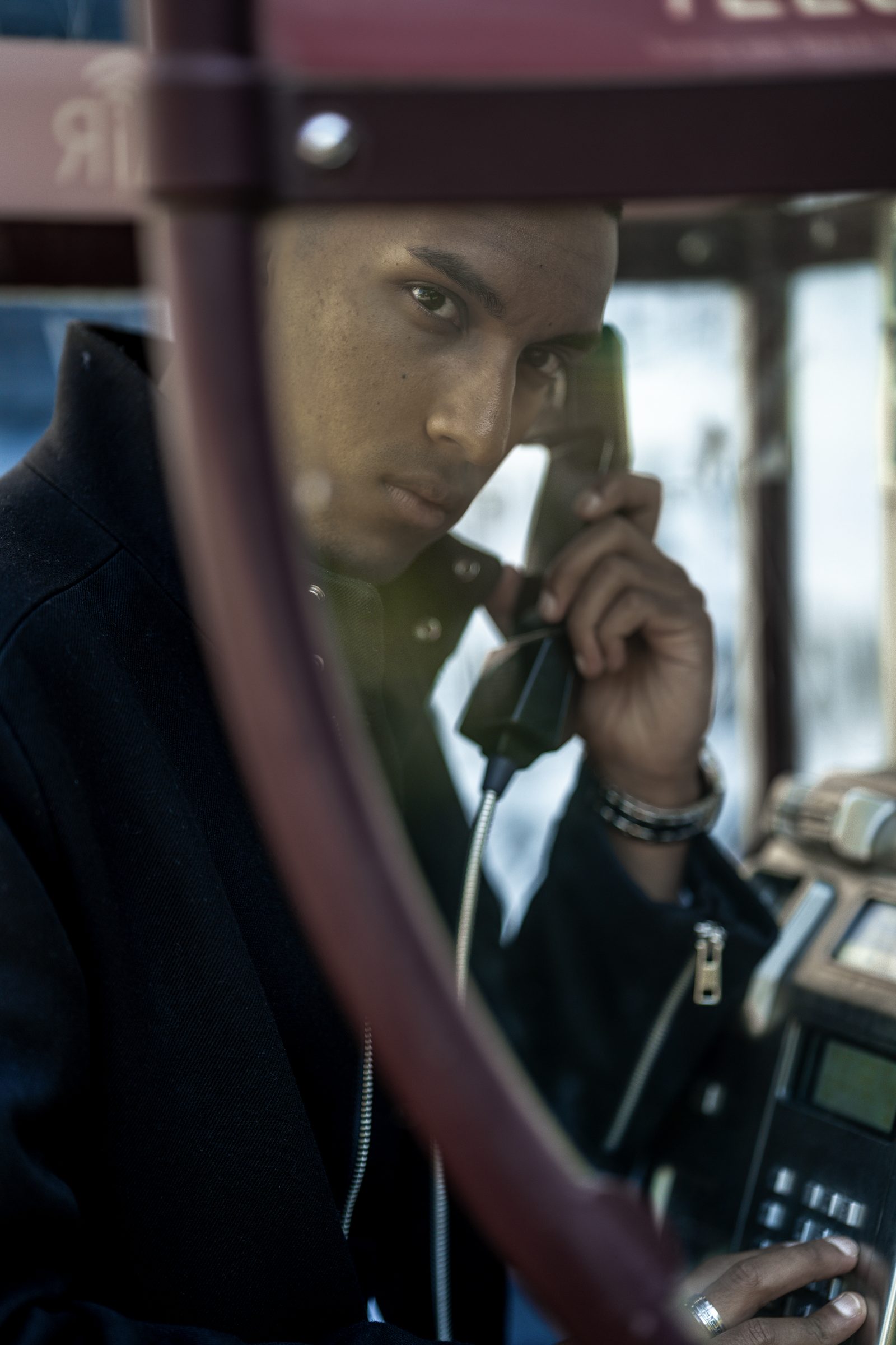 A man holds a payphone handset inside a red telephone box, seen through the curved glass of the door