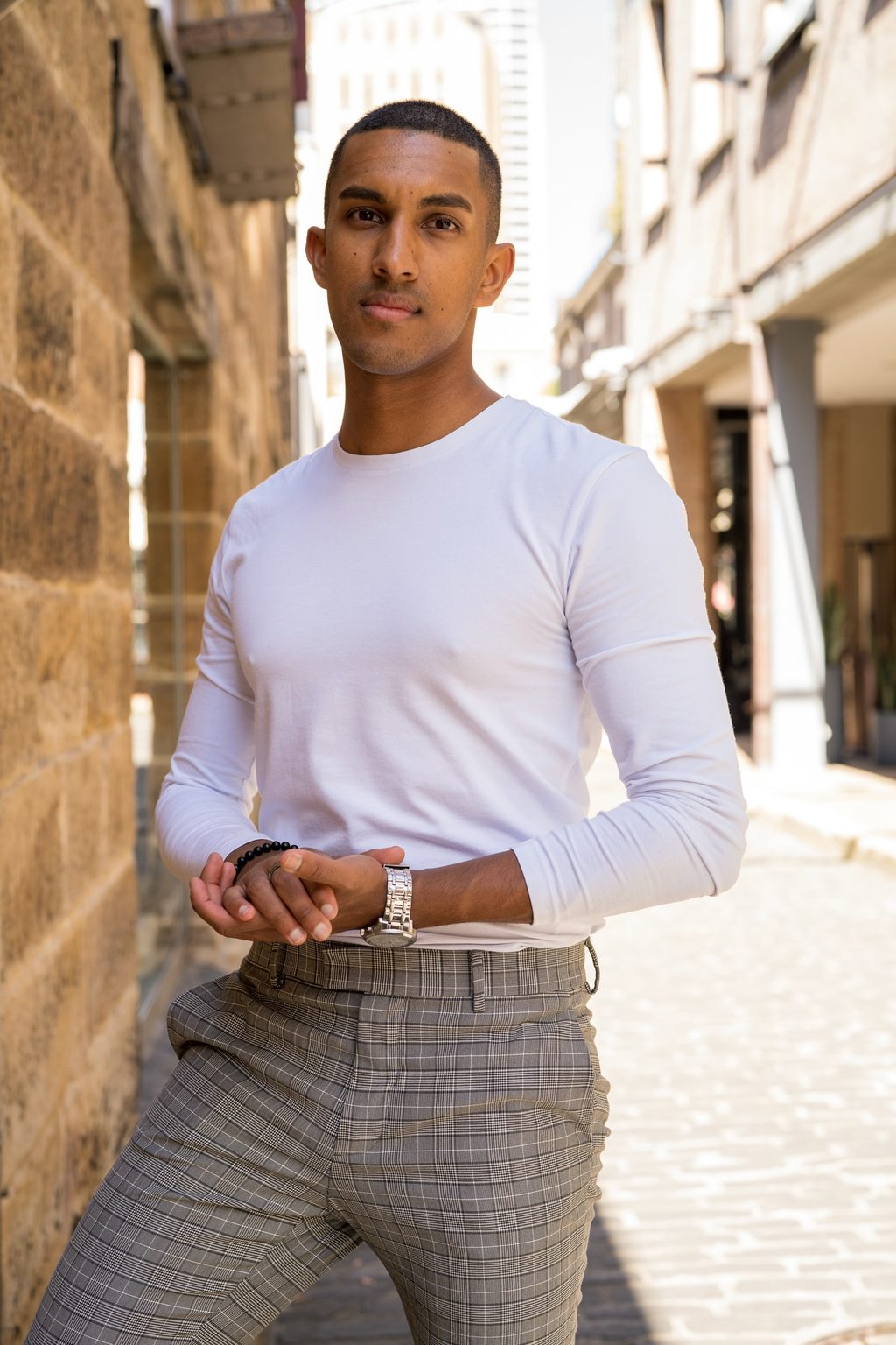 A man in a white long-sleeve top and checked trousers stands with hands clasped in a sunlit sandstone laneway between old buildings