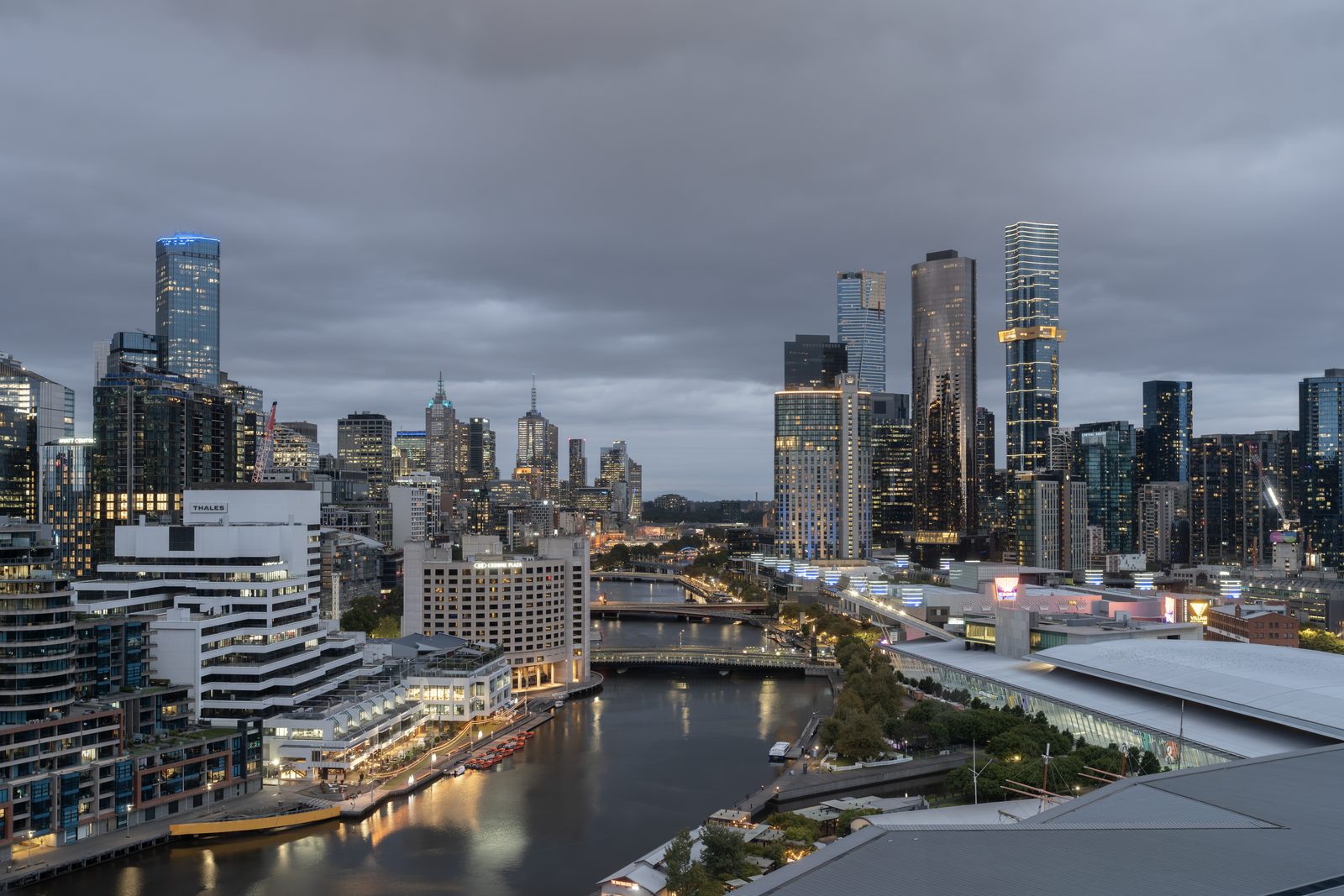 Melbourne's towers stand under heavy grey cloud at dusk, the Yarra running dark between lit riverside buildings.