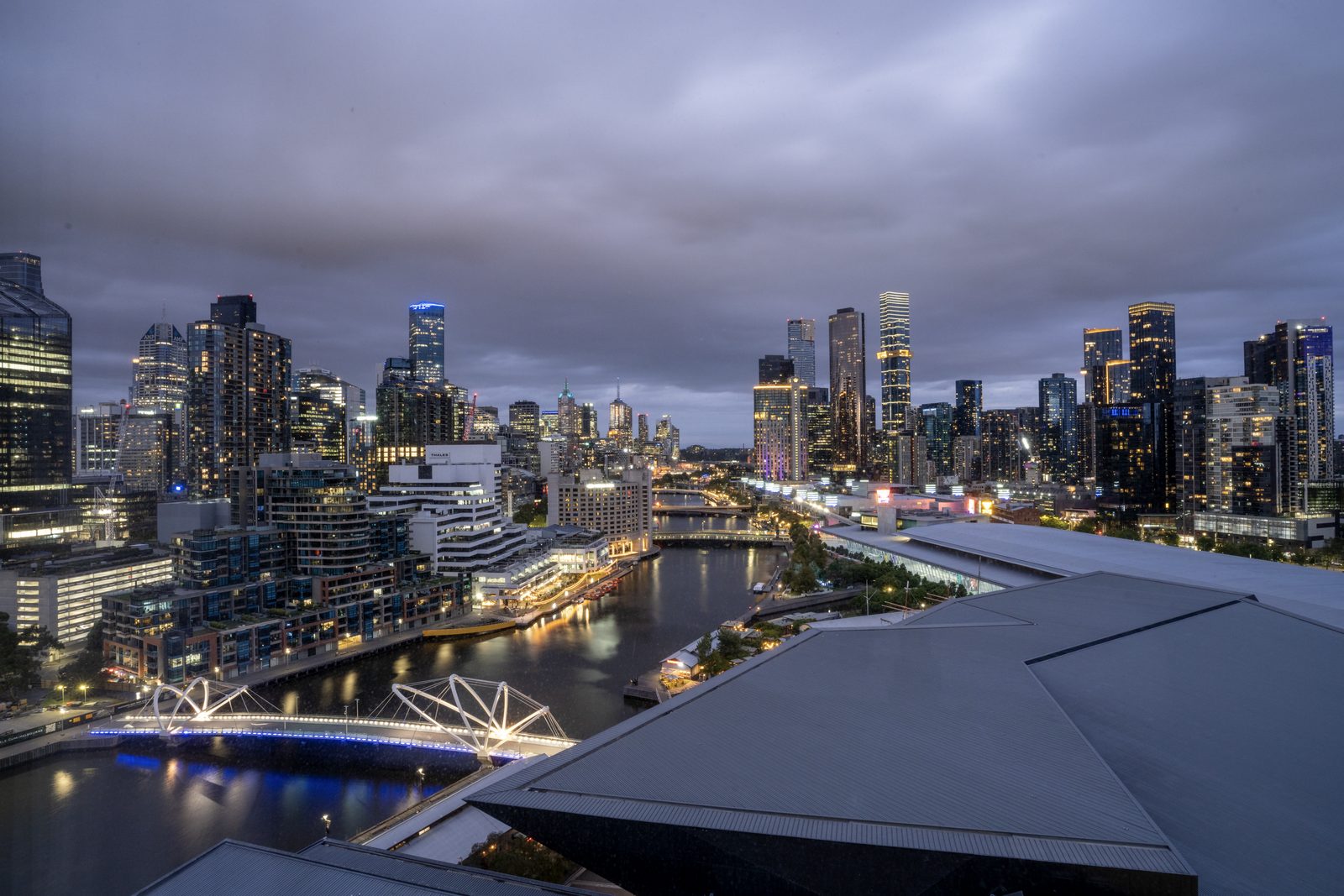 A lit pedestrian bridge crosses the Yarra below Melbourne's towers at dusk, pale roofs angling across the foreground.