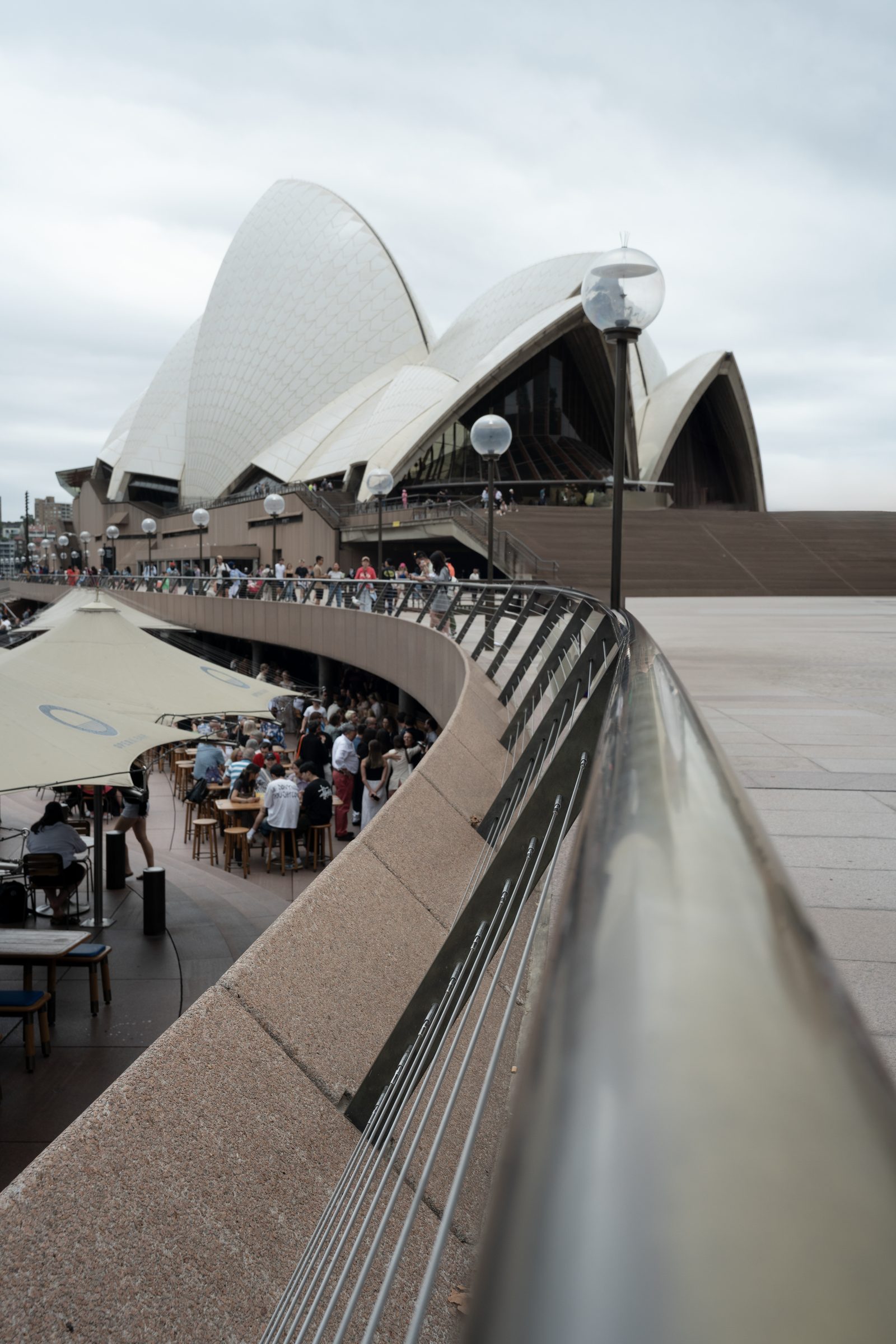 A polished steel handrail curves toward the tiled shells of the Sydney Opera House, umbrellas and crowded tables below.