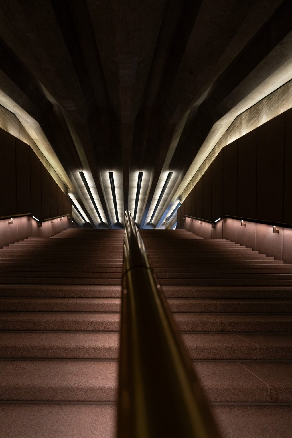 A long stone stairway descends beneath a fan of angled concrete beams lit by rows of recessed lights.