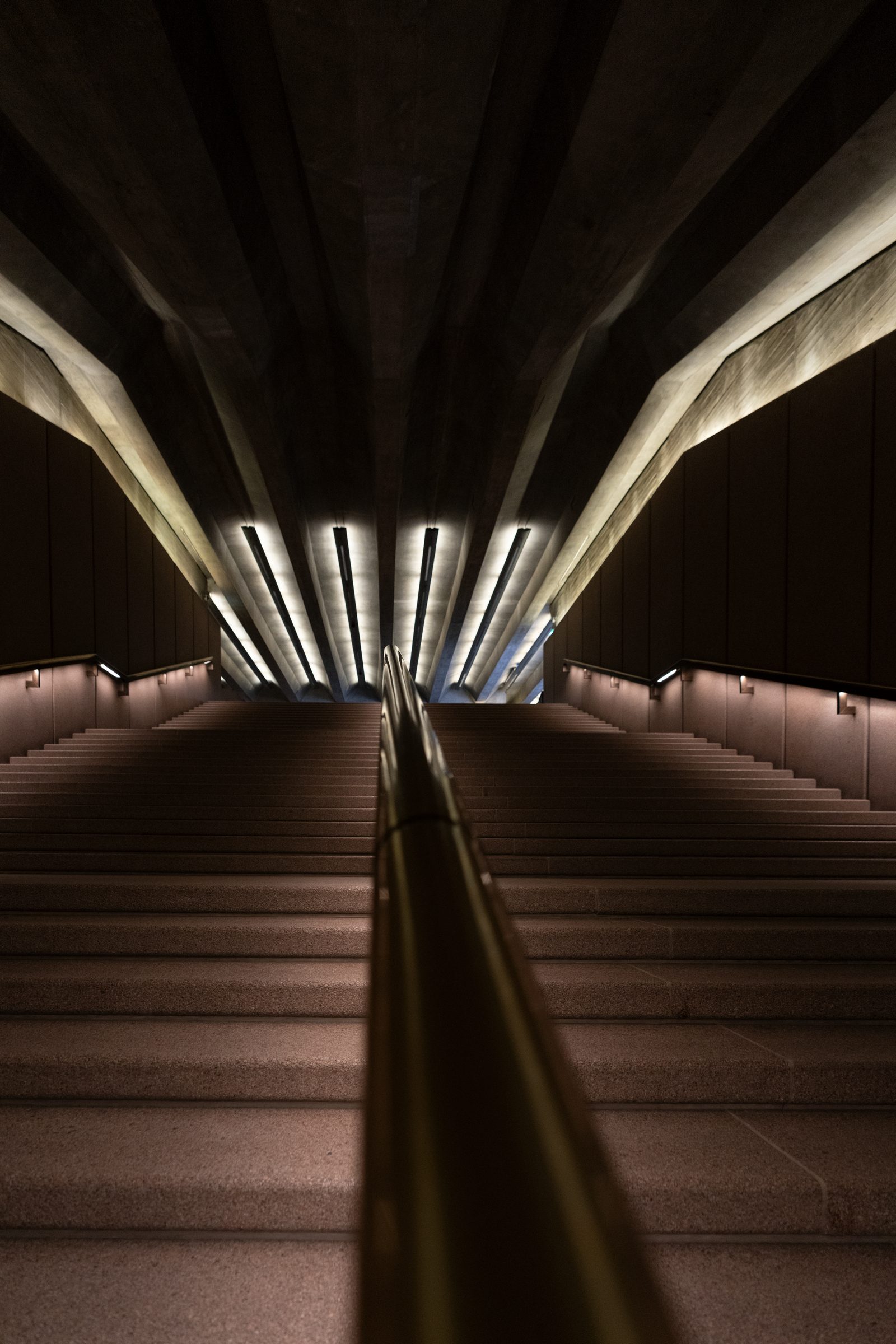 A long stone stairway descends beneath a fan of angled concrete beams lit by rows of recessed lights.