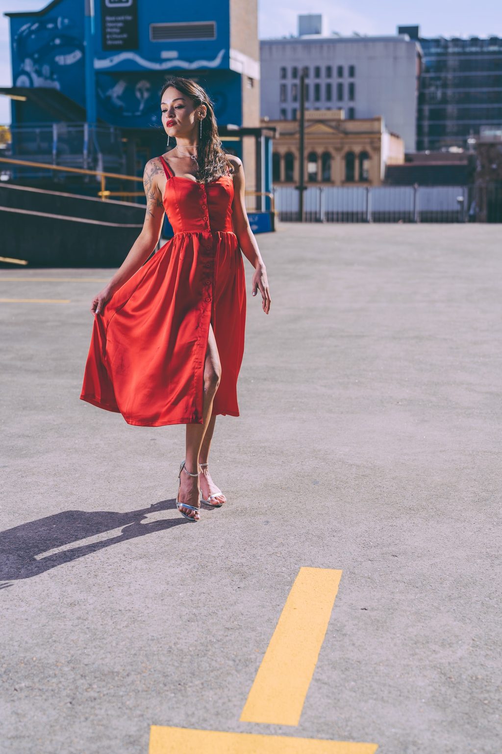 A woman in a flowing red satin dress and silver heels walks across an open rooftop car park with city buildings in the distance