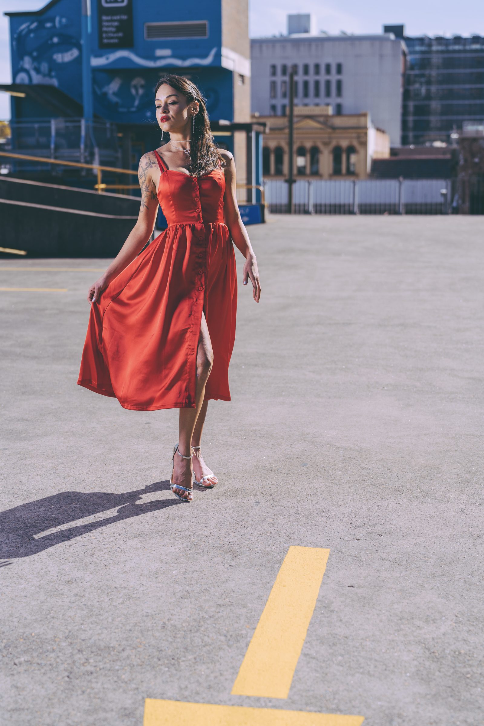 A woman in a flowing red satin dress and silver heels walks across an open rooftop car park with city buildings in the distance