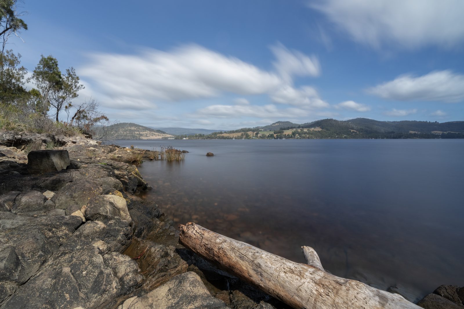 A bleached log lies on a rocky shore beside still water, long-exposure cloud streaking above the low hills opposite.