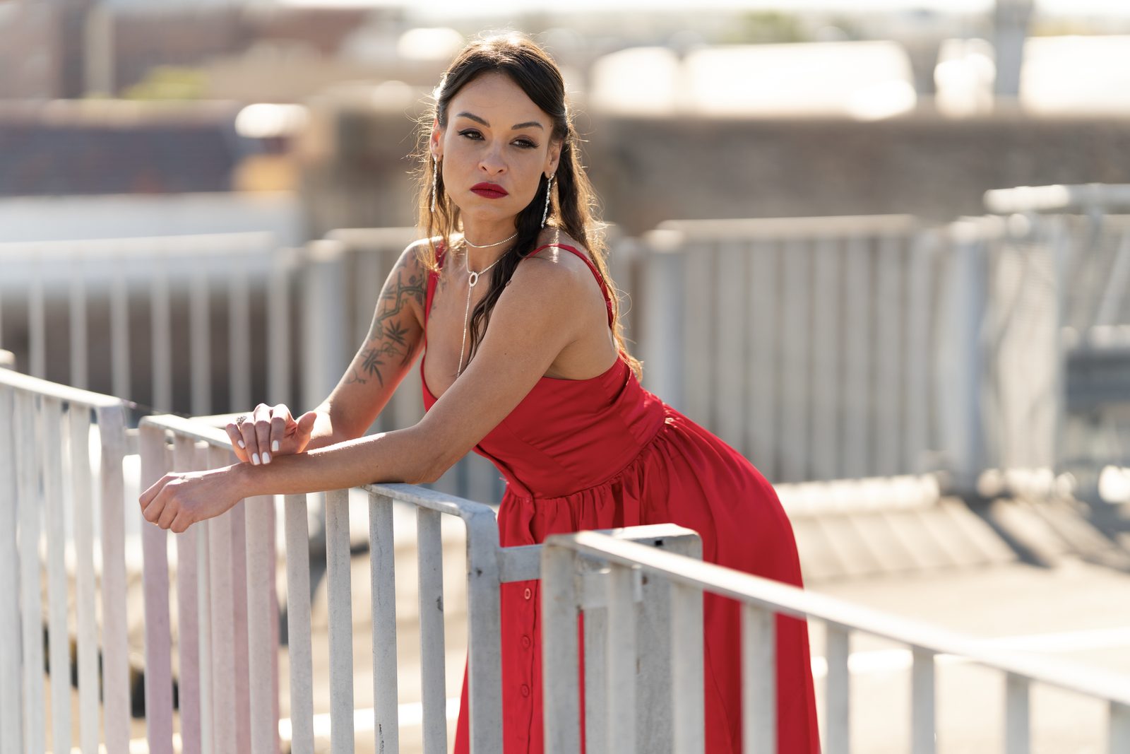 A woman in a red halter dress leans both arms on a white rooftop railing, city rooftops soft behind her