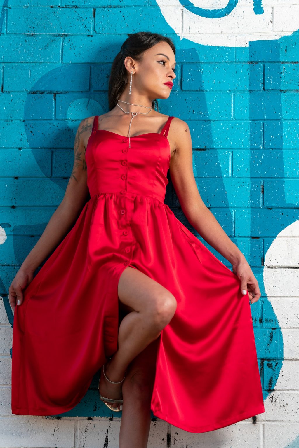 A woman in a red satin button-front dress leans against a teal painted brick wall, one leg raised and gaze turned aside