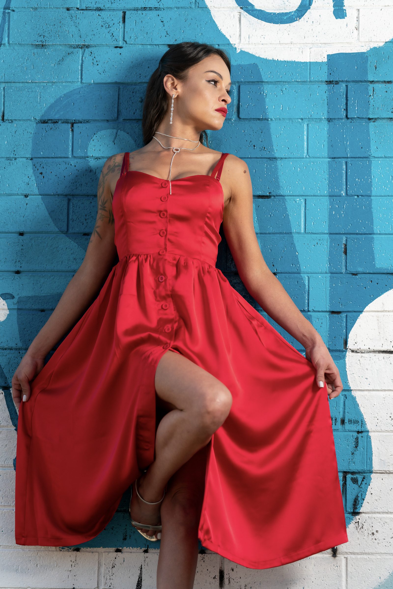 A woman in a red satin button-front dress leans against a teal painted brick wall, one leg raised and gaze turned aside