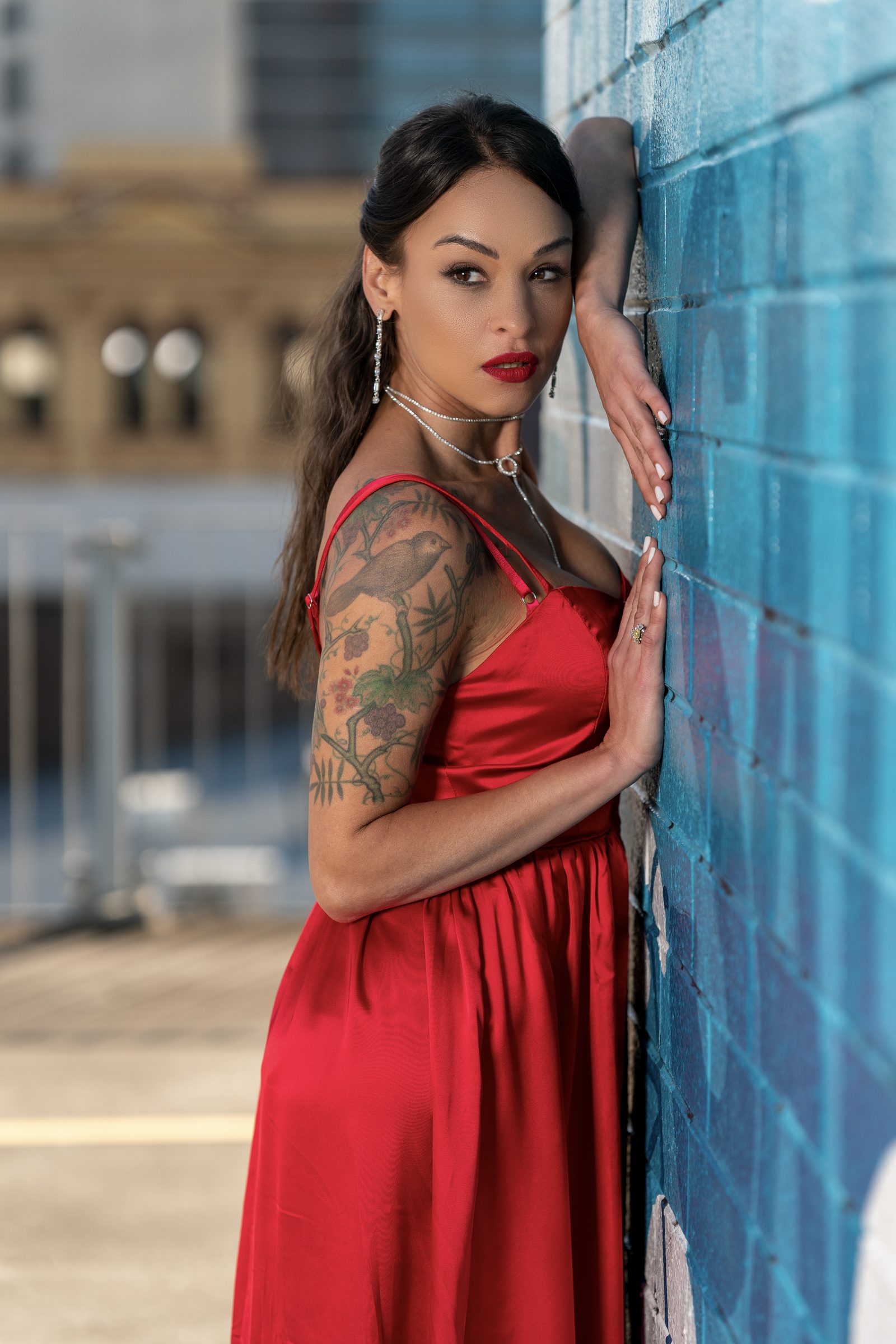 A woman in a red satin dress leans against a blue painted brick wall with both hands raised to it, a tattoo sleeve on her arm
