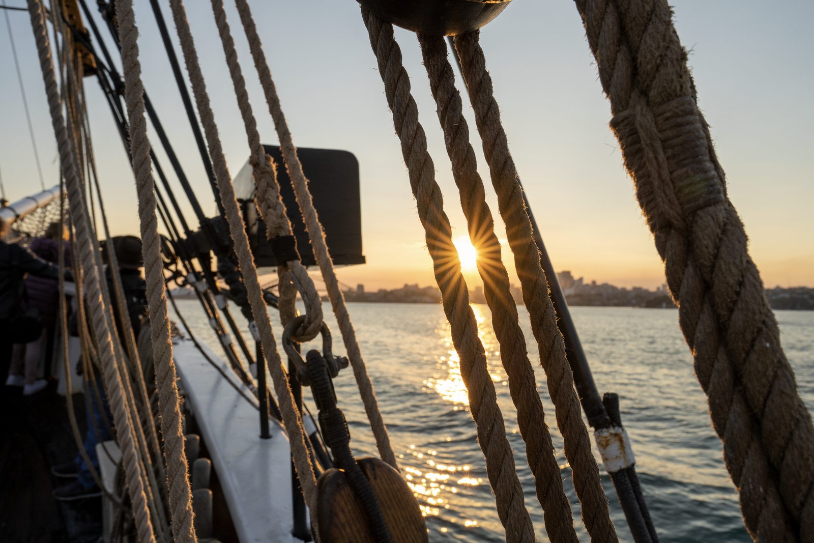 The sun bursts between the ropes of a tall ship's rigging at sunset, its light laid in a path across the harbour.