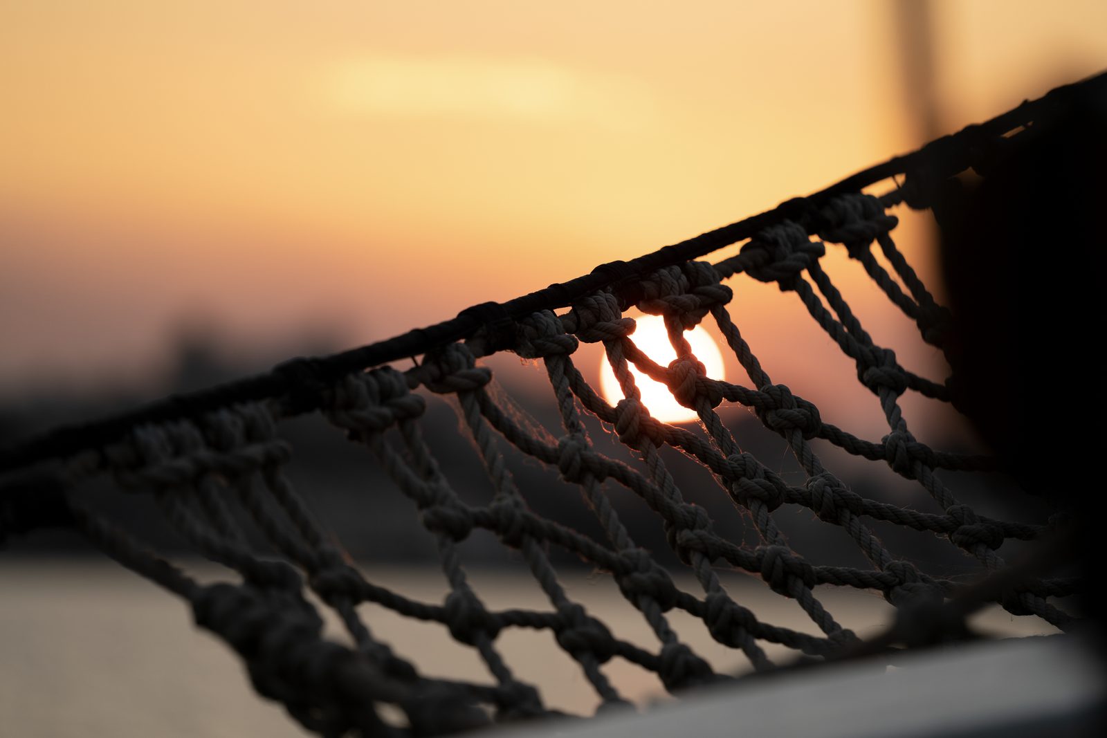 Knotted rope netting is silhouetted against an orange sunset, the sun sitting in one gap of the mesh.