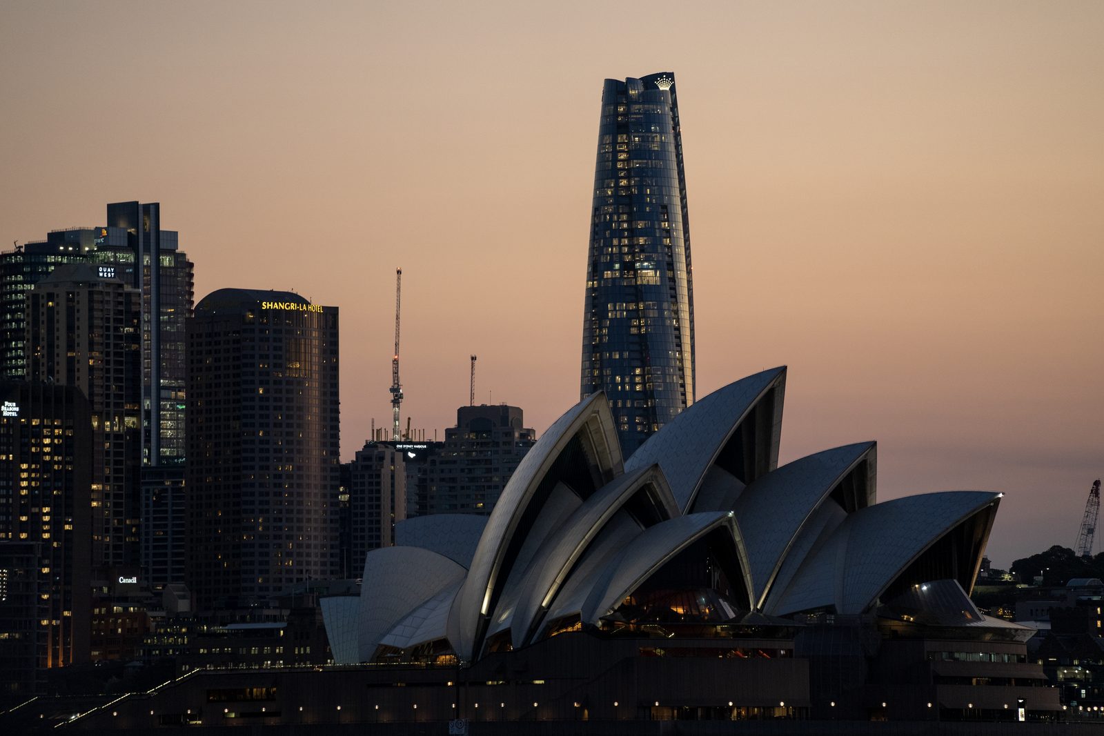 The Sydney Opera House shells sit below city towers against a warm dusk sky, harbour lights just coming on.
