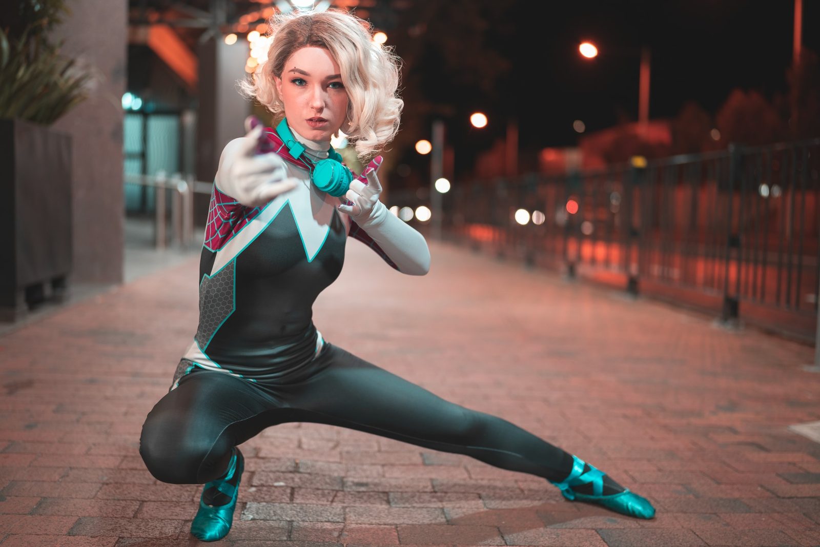 A cosplayer in a teal, white and pink spider-themed costume crouches on a night street, one hand reaching toward the camera