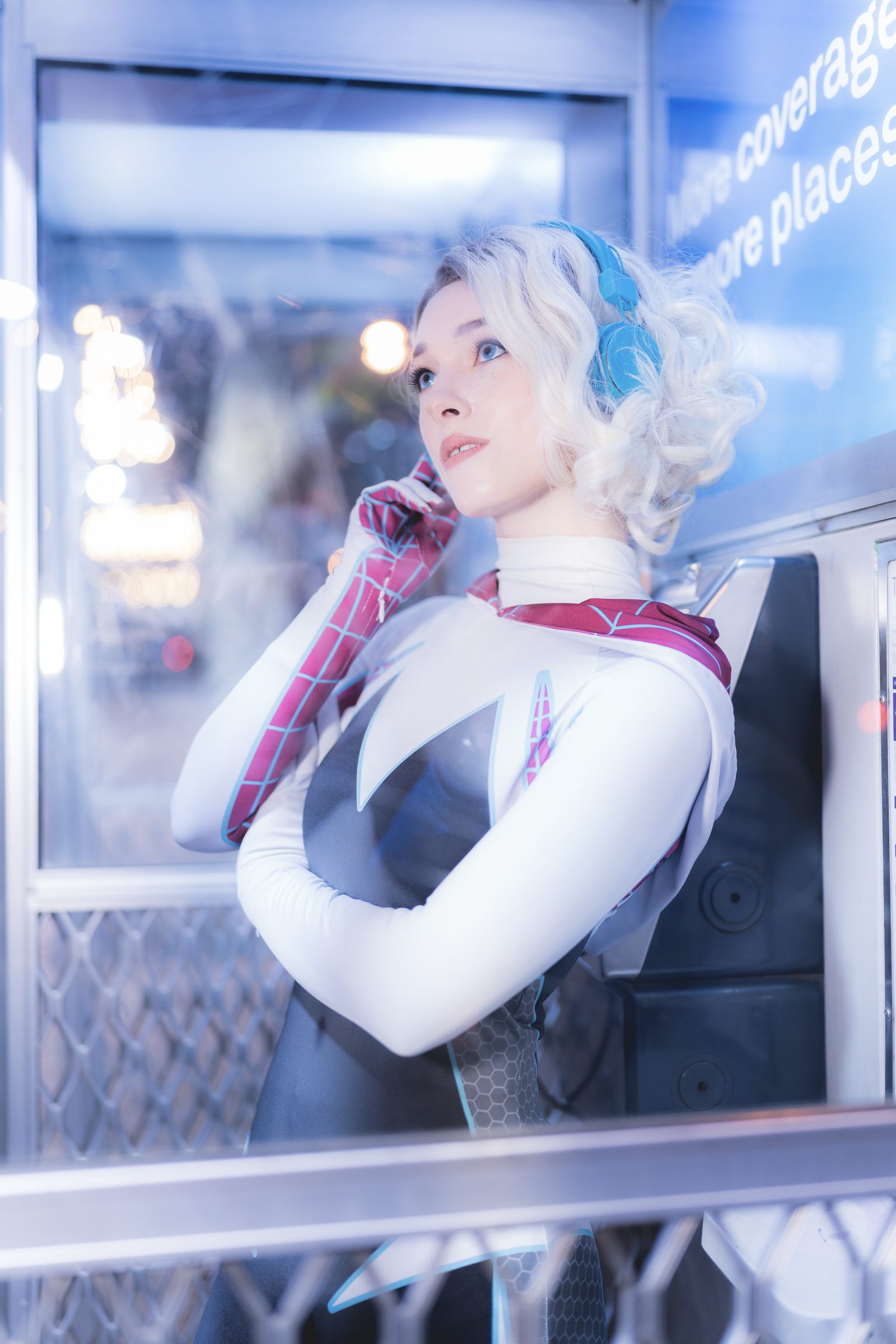 A cosplayer in a white and pink spider-themed suit with teal headphones leans against a lit phone booth, looking up