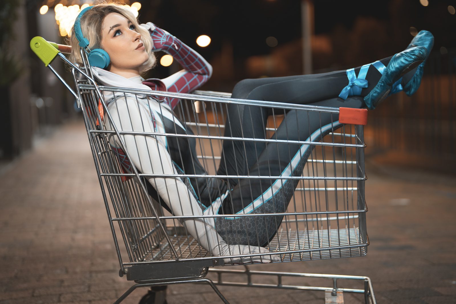 A cosplayer in a hooded spider suit and teal headphones reclines in a shopping trolley on a paved street at night